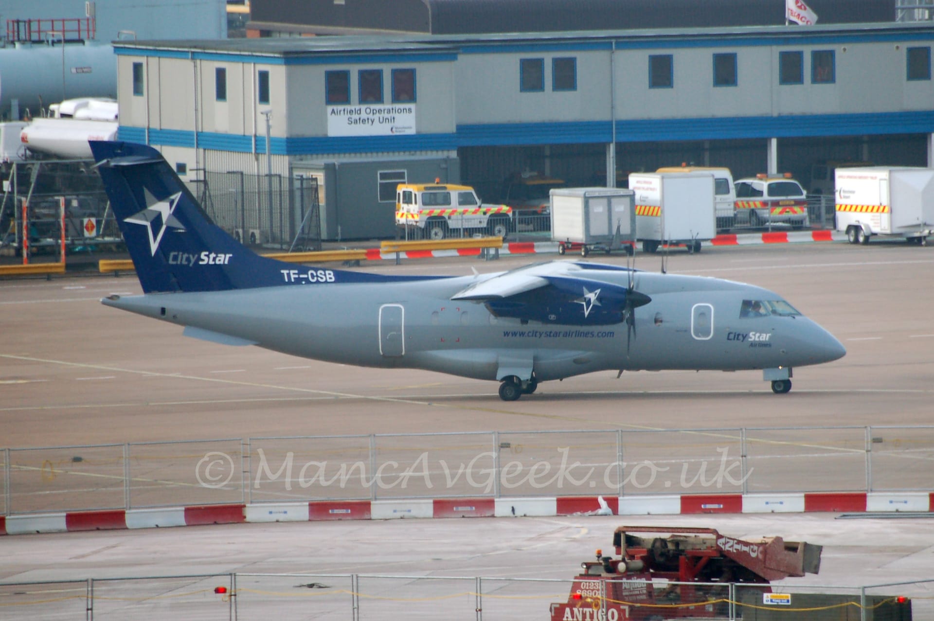 Side view of a high-winged, twin propellor-engined airliner taxiing from left to right. The planes body is grey, with dark blue text "www[dot]CityStarAirlines[dot]com"on the lower fuselage below the wings, and larger "City Star Airlines" under the cockpit.. The engine pods are dark blue, with white stars towards the front. The tail is the same dark blue, with a larger grey and white star, with grey and white "City Star" titles underneath. Grey concrete apron fills the foreground, with part of it partitioned off by metal fences set into red and white concrete blocks. A grey, 2 storey building with a white "Airfield operations Safety Unit" sign stretches across the frame from the right, with white vehicles with a banded orange and yellow stripe running along the body.