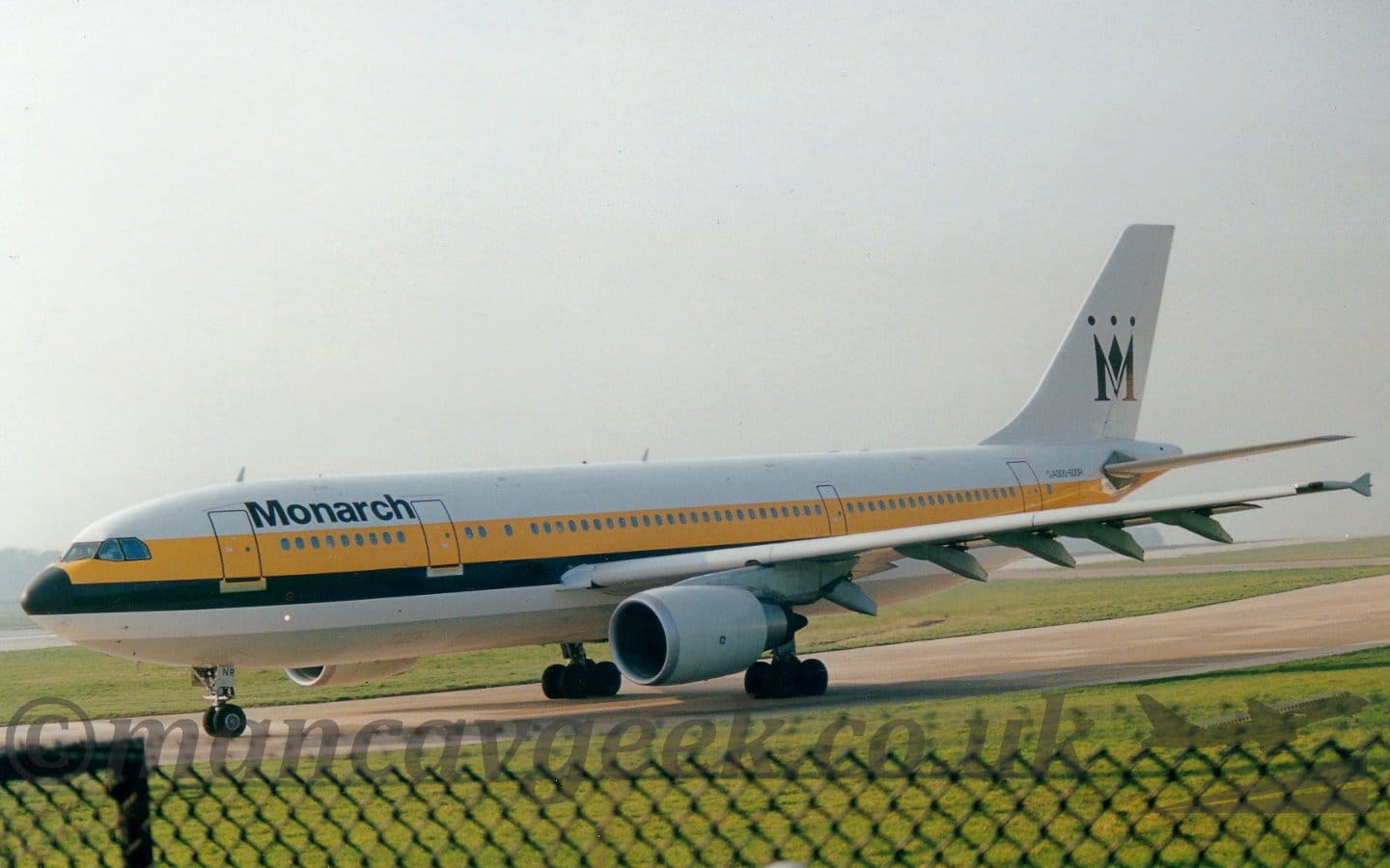 Side view of a white, twin engined jet airliner taxiing from right to left. The plane has a thick yellow and black stripe running along the body, covering the passenger cabin windows, and black "Monarch" titles on the upper forward fuselage. There is a large, stylised, black crown on the white tail, resembling an upper-case letter "M". The top of a black chain link fence stretches across the bottom of the frame, with green grass filling the rest of the foreground. Bright but hazy white sky fills the rest of the frame.