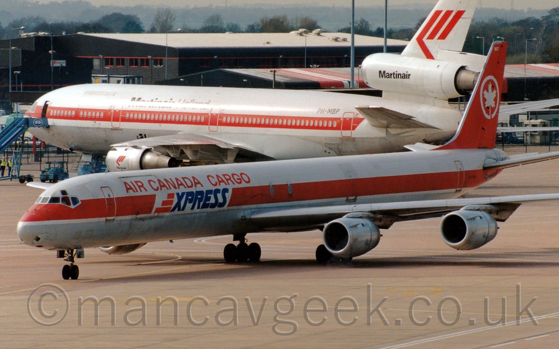 Side view of a long and thin, white and red, 4 jet-engined cargo plane taxiing from right to left. The plane is mostly white, with a grey belly, and a thick red stripe running along the body. There are red "Air Canada Cargo" titles on the upper forward fuselage, with blue "Express" titles just below. The tail is red, with the white outline of a circle, with a white maple leaf in the middle. Grey concrete apron fills the foreground. A white, 3 engined jet airliner with a red stripe running along the body and black "MartinAir" titles on the centre engine is in the background, with black buildings beyond that, under grey skies.
