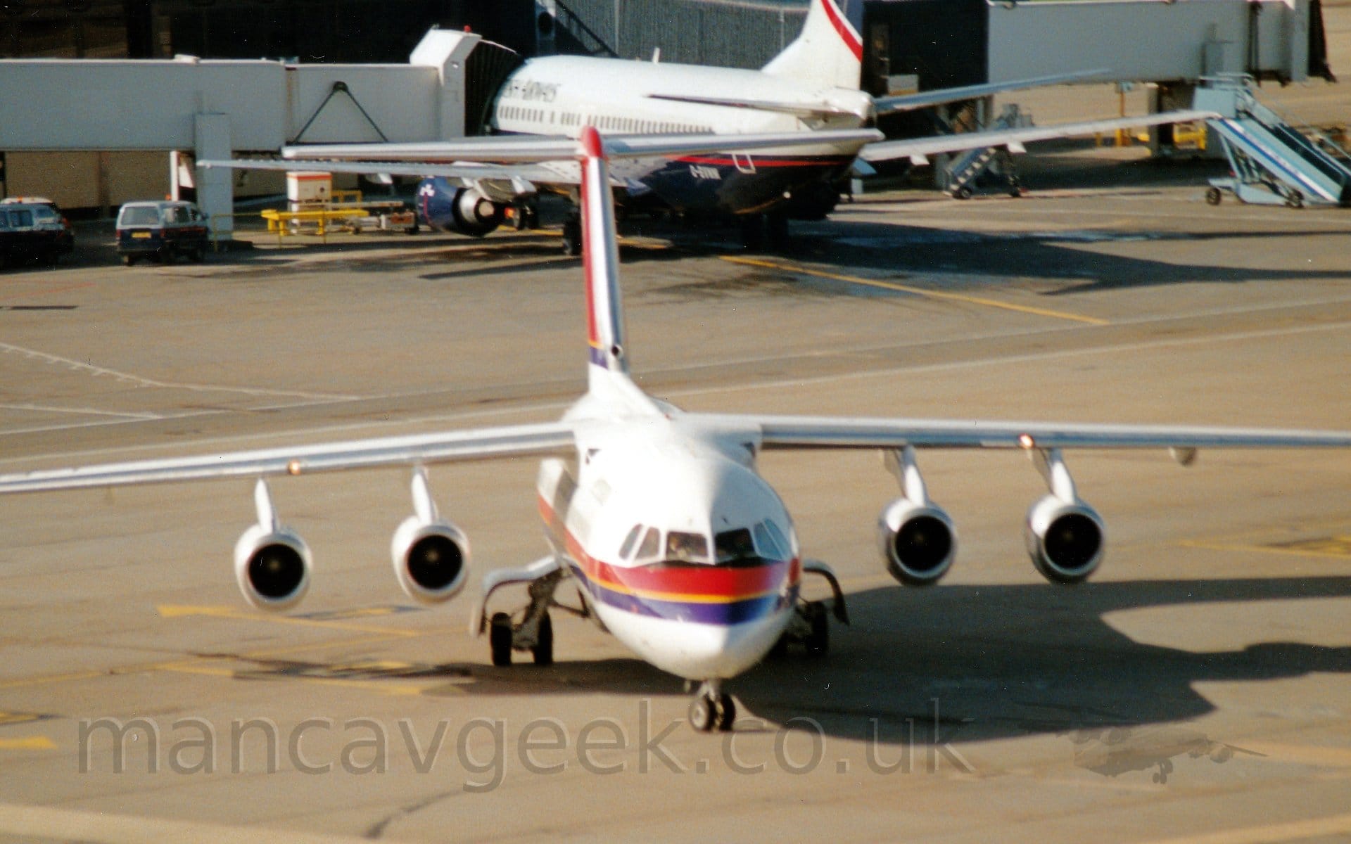 Forward view of a high-winged, 4 engined jet airliner taxiing from left to right, almost directly towards the camera. The plane is white, with a red and purple stripe with yellow piping in the middle running along the body, and a red tail with a purple stripe with yellow piping at the bottom. It's nose wheel is turned to it's left, as it turns to the right of the frame. In the background, a grey and dark blue, twin engined jet airliner facing away from the camera, has an airbridge connected to it's forward fuselage, the other end of the bridge connected to a grey building.