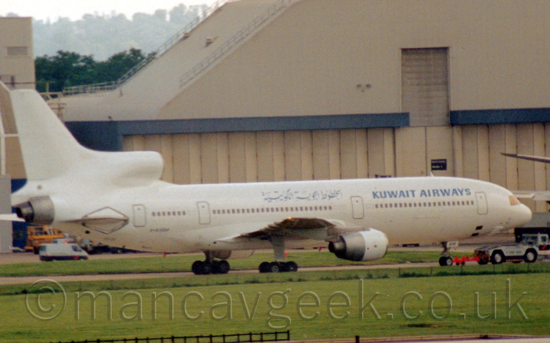 Side view of a white, 3 engined jet airliner being towed from left to right by a low-slung, grey and blue truck. The plane is almost entirely white, with blue "Kuwait Airways" titles on the upper fuselage, in English towards the front, and in an Arabic script over the wings. Green grass fills the foreground, with a large cream-coloured hangar dominating the background.