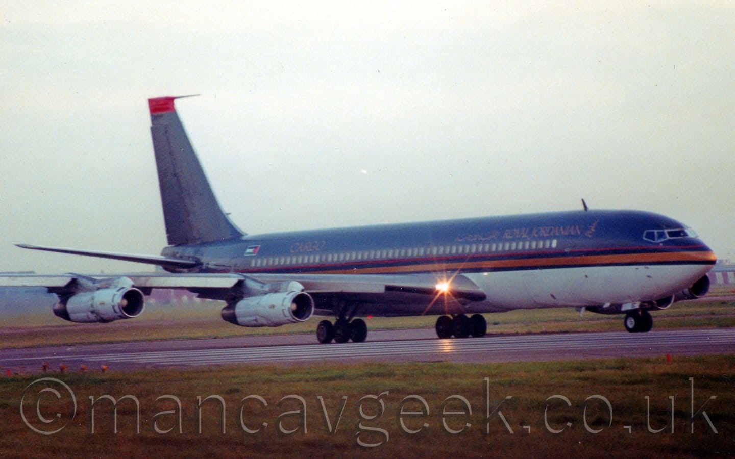 Side view of a dark purple, 4 engined jet airliner taxiing from left to right. The plane has a grey belly, with a golden stripe and red piping running along the body. There are gold "Royal Jordanian" titles on the upper forward fuselage, in English to the front, repeated in an Arabic script to the rear, with additional "golden "Cargo" titles on the upper rear fuselage, just ahead of a Jordanian flag. Green grass fills the foreground, with more grass visible under the plane in the background, with flat grey sky filling the rest of the frame.