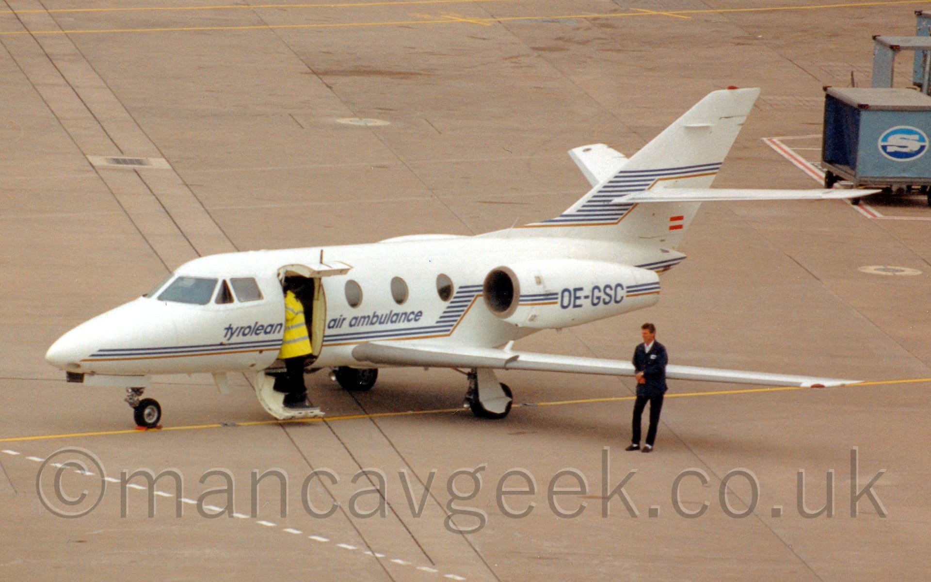 Side view of a twin engined Bizjet with the engines mounted on the sides of the rear fuselage, parked on grey concrete apron facing to the left. The plane has blue and brown piping running along the planes body and engine pods, as well as along the tail. The registration "OE-GSC" is on the engine pods in blue. A man wearing a blue coat and black pants is leaning on the planes left wing, while another man wearing a hi-viz coat is standing on the planes steps (actually the lower half of the planes door, folded downward, the upper half folded upwards), leaning in to the plane. Grey concrete apron fills the rest of the frame.