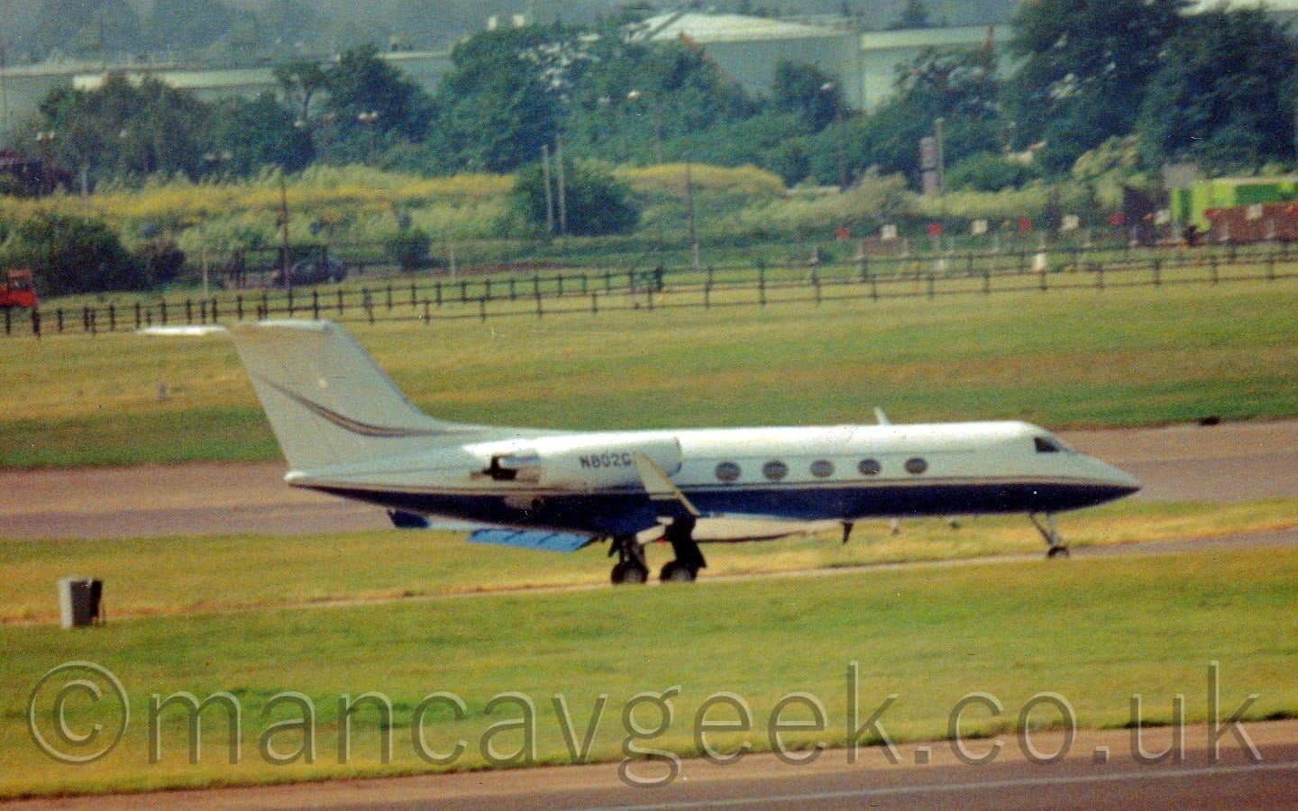 Slightly blurry and grainy, side view of a twin engined BizJet taxiing from left to right. The plane is mostly white, with a dark blue belly, and a yellow stripe running along the body, with another yellow stripe on the tail. A black runway bordered by large areas of grass fills the foreground, with more grass leading up to trees in the background.