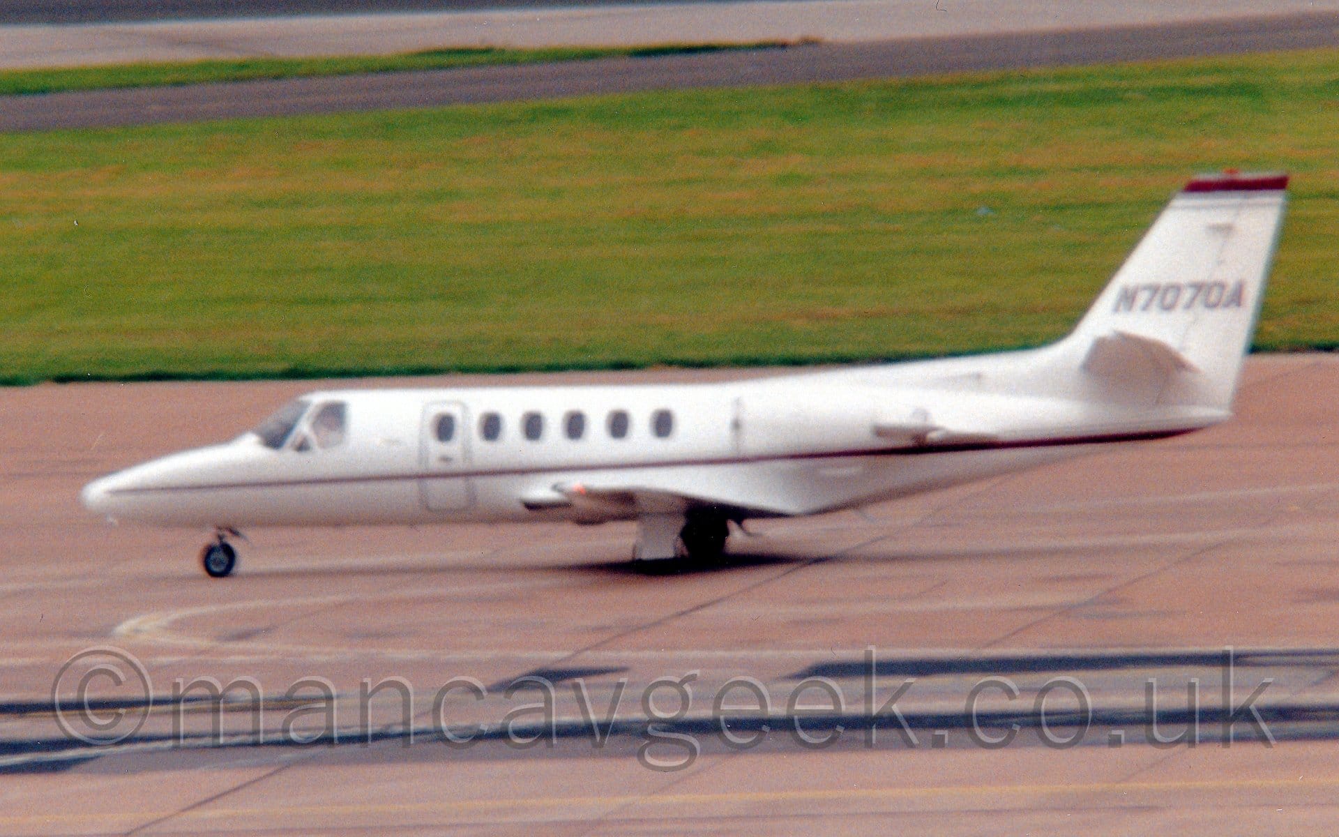 Slightly blurry side view of a white, twin engined BizJet, taxiing from right to left. The plane has a thin red and grey stripe running along the body from the nose, getting slightly wider as it moves rearwards. A slightly thicker version of this stripe runs along the top of the tail. The grey registration "N7070A" is on the tail. Grey concrete apron fills the foreground, with a large area of green grass in the background leading up to another taxiway and a runway in the distance.