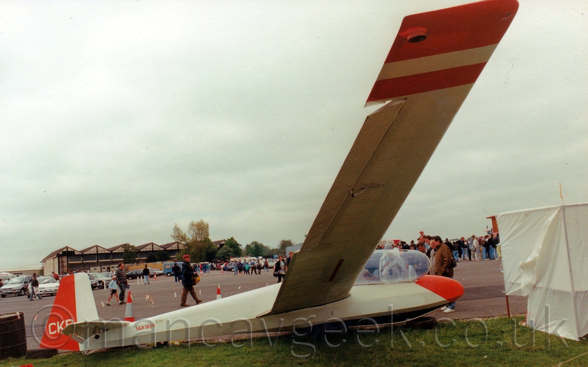 Side view of a cream-coloured glider parked on grass facing to the right, it's left wingtip on the ground and it's right wingtip up in the air. The nose and the rear half of the tail are bright red, as are the wing-tips. The small, red registration "BGA-1601" is on the upper rear fuselage, just in front of the tail, with cream letters "CKR" on the red portion of the tail. The canopy on the tandem 2 seat cockpit is open, hinged to the right. Crowds of people can be seen walking past in the background, with rows of parked cars and black and white hangars in the distance, under flat grey skies.