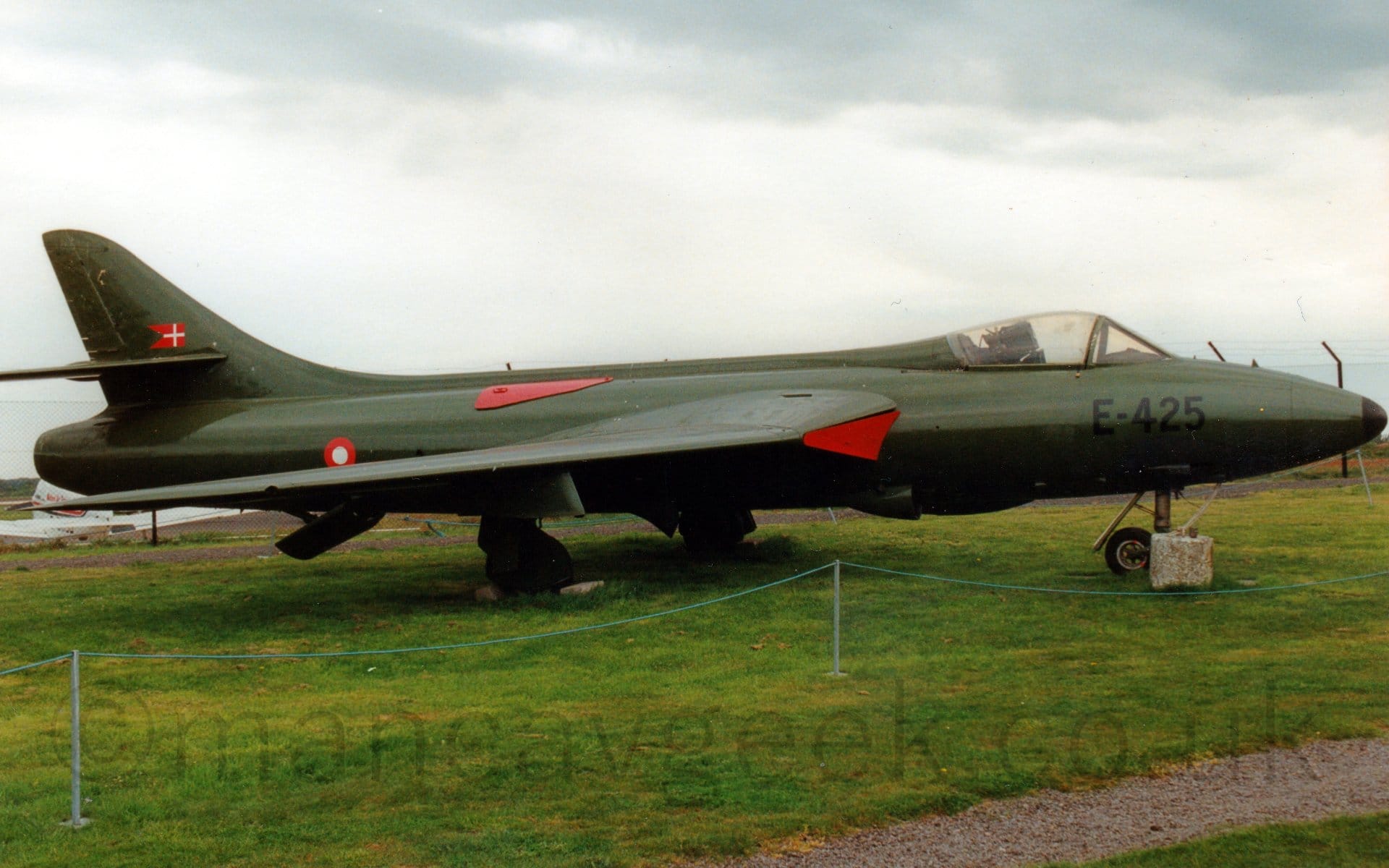 Side view of an olive green, single engined military fighter jet aircraft, parked on grass facing to the right. The black serial "E-425" is is on the plane's nose, just under the cockpit bubble. There is a red and white roundel on the rear fuselage, with a small red pennant with a white cross in the middle on the tail. There are red covers on the air intakes in the wing root, with a red metal cover on the upper rear fuselage. Green grass fills the bottom of the frame, with grey skies filling the top of the frame.