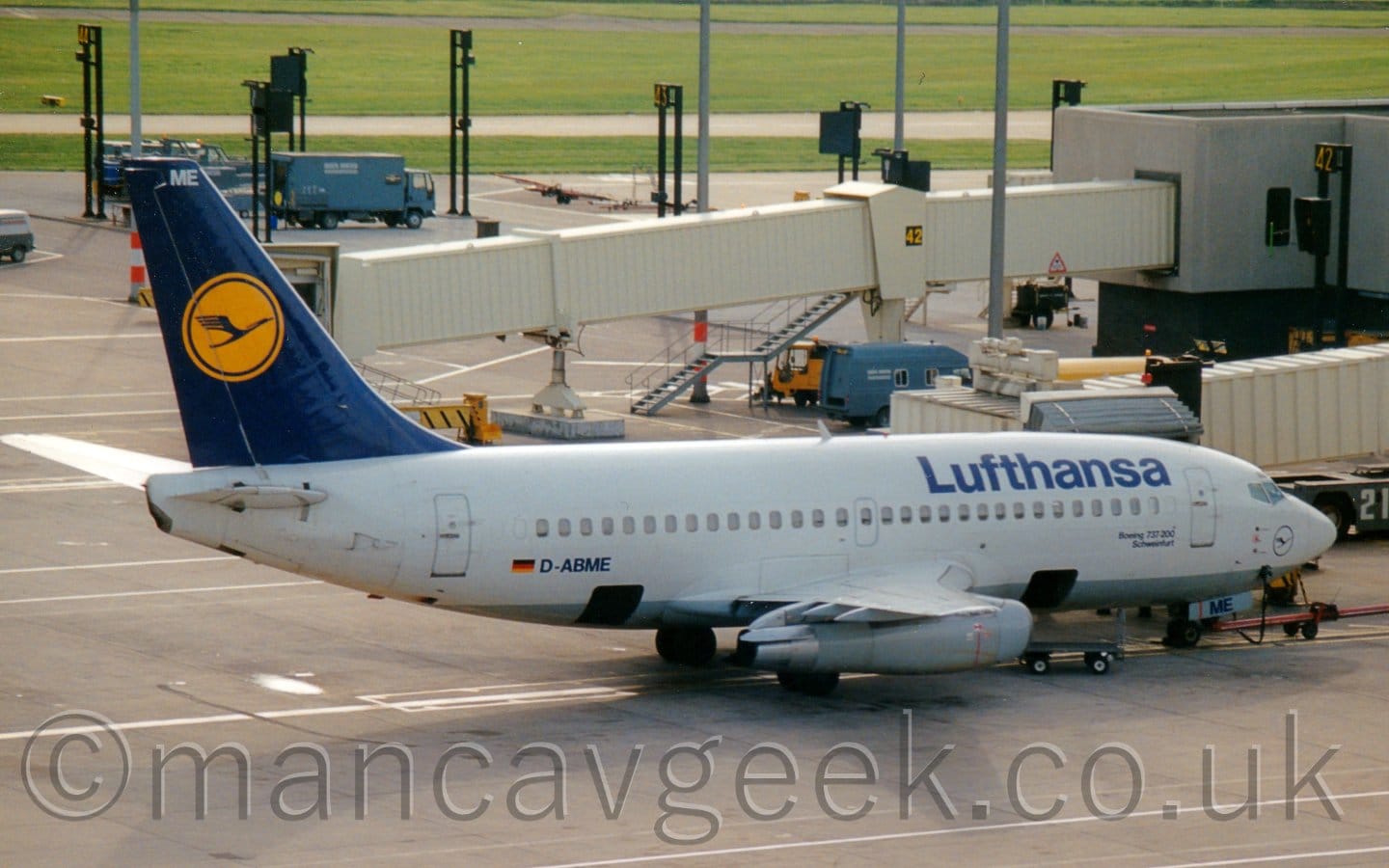 Side view of a white, twin-engined jet airliner parked facing to the right. The plane has a grey belly, and dark blue "Lufthansa" on the upper forward fuselage. The blue registration "D-ABME" is on the lower rear fuselage, next to a small German flag. The tail is the same dark blue, with a yellow circle containing a stylised image of a blue flying bird. Grey concrete apron fills the foreground, with a pair of cream-coloured airbridges stretching across the frame, attached to a terminal building on the right of the image. Tall black gate markers stretch towards the top left of the frame from the terminal, with a blue truck parked amongst them. Green grass beyond that stretches off into the distance.