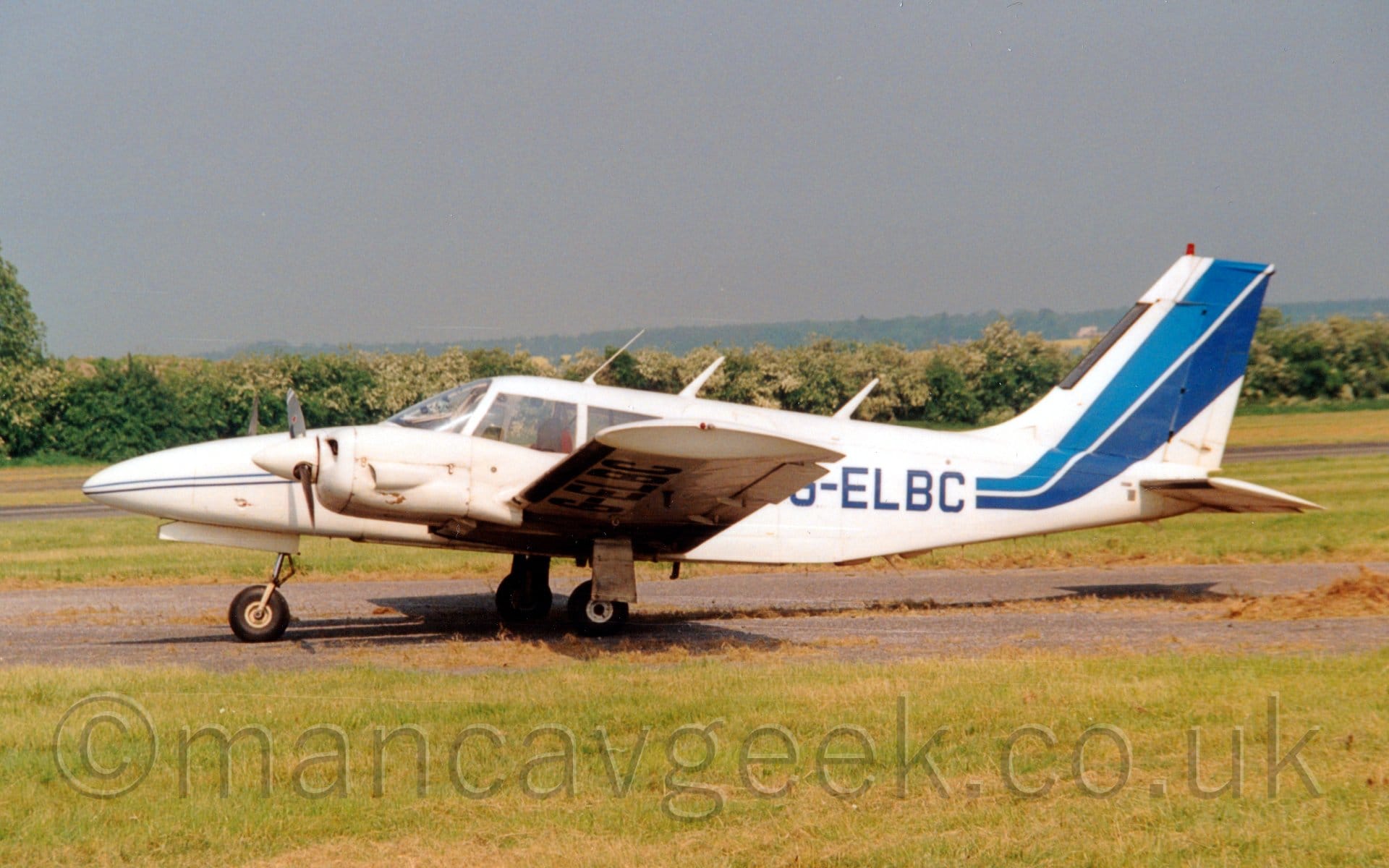 Side view of a white, twin propellor-engined light aircraft parked on tarmac facing to the left. The plane is mostly white, with a pair of thin stripes, one light blue, the other dark blue, running along the body, getting wider as the move rearwards, until they sweep up to fill most of the tail. The dark blue registration is on the rear fuselage, as well as under the left wing. Green grass fills the foreground, with trees stretching across the background, under grey skies.