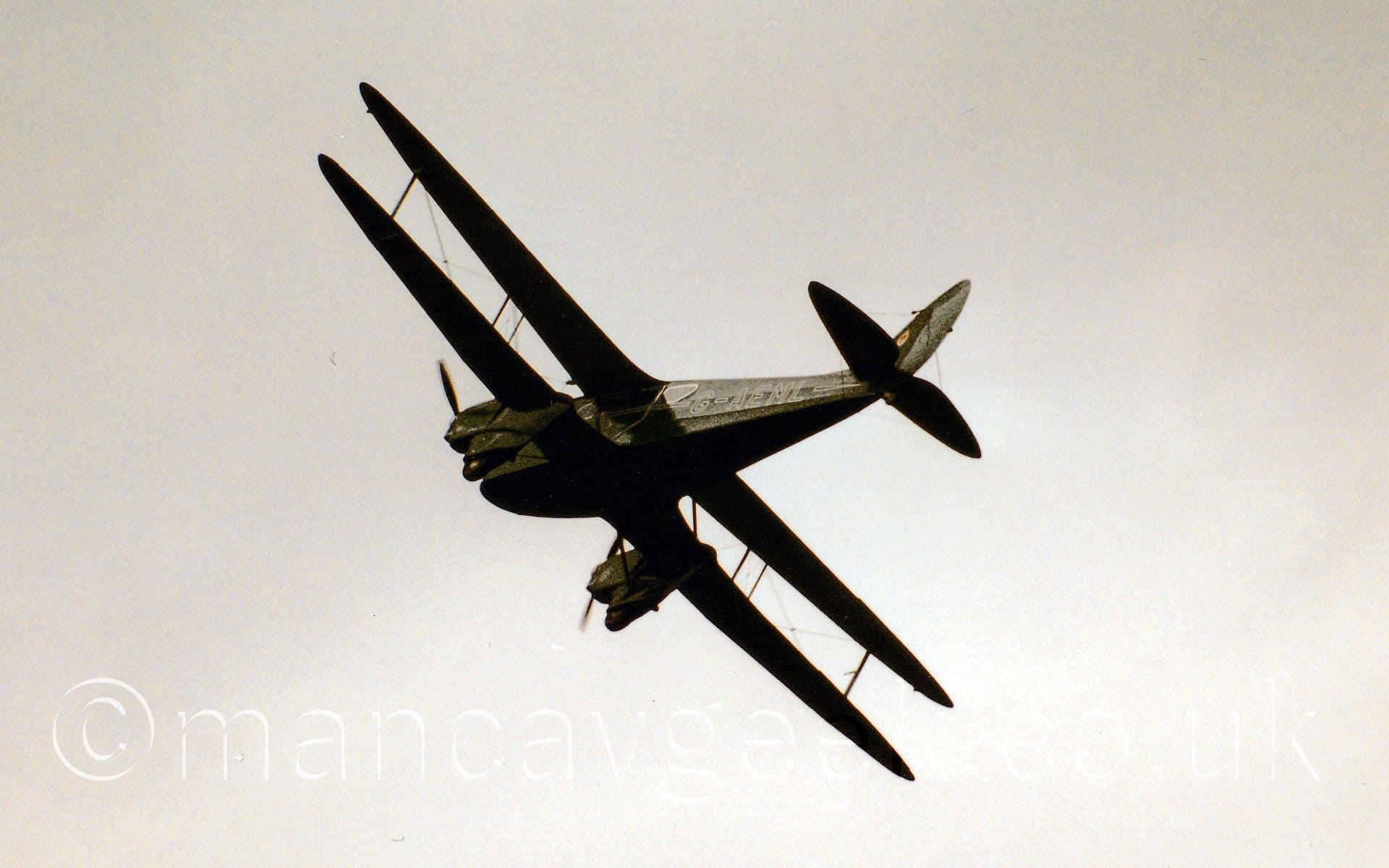Low side view of a green, twin engined bi-plane airliner flying from right to left at a low altitude, banking to the right and flying away from the camera. The registration "G-AEML" is on the side of the rear fuselage, outlined in white. Flat white sky fills the rest of the frame.