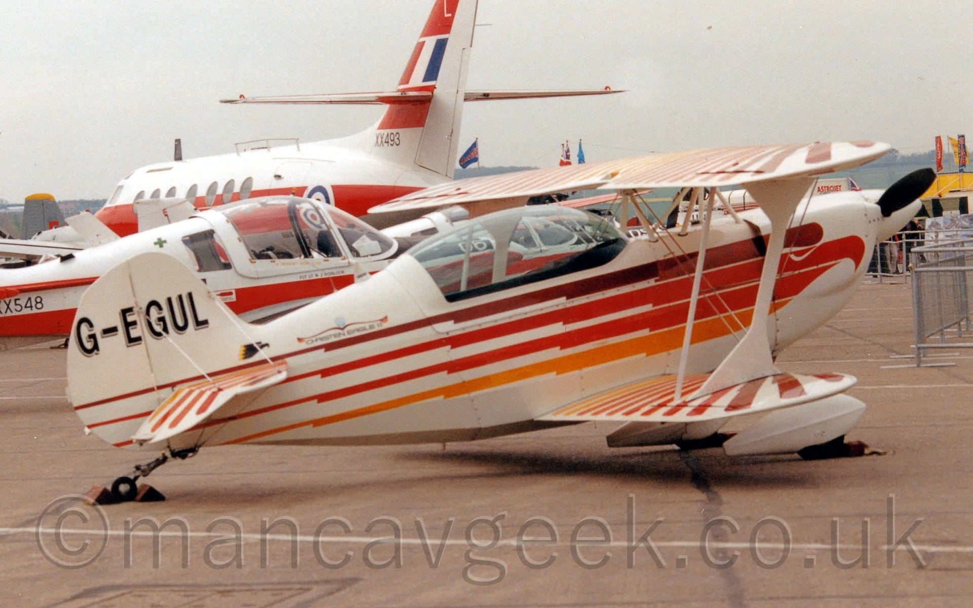 Side view of a white, single engined bi-plane light aircraft parked facing to the right. There are brown, orange, red, and yellow stripes running along the body, forming a stylised image of the head and body of an eagle, with the eagles wings on both the top and bottom of the wings. The black registration "G-EGUL" is on the tail, with a grey drop shadow. Grey concrete apron fills the foreground. In the background, a couple of white and red planesd can be seen, one is a red and white military trainer with the serial "XX548" on the rear fuselage, the other being a twin propellor-engined airliner in a similar livery and the serial "XX493" on the tail. Grey sky fills the rest of the frame.