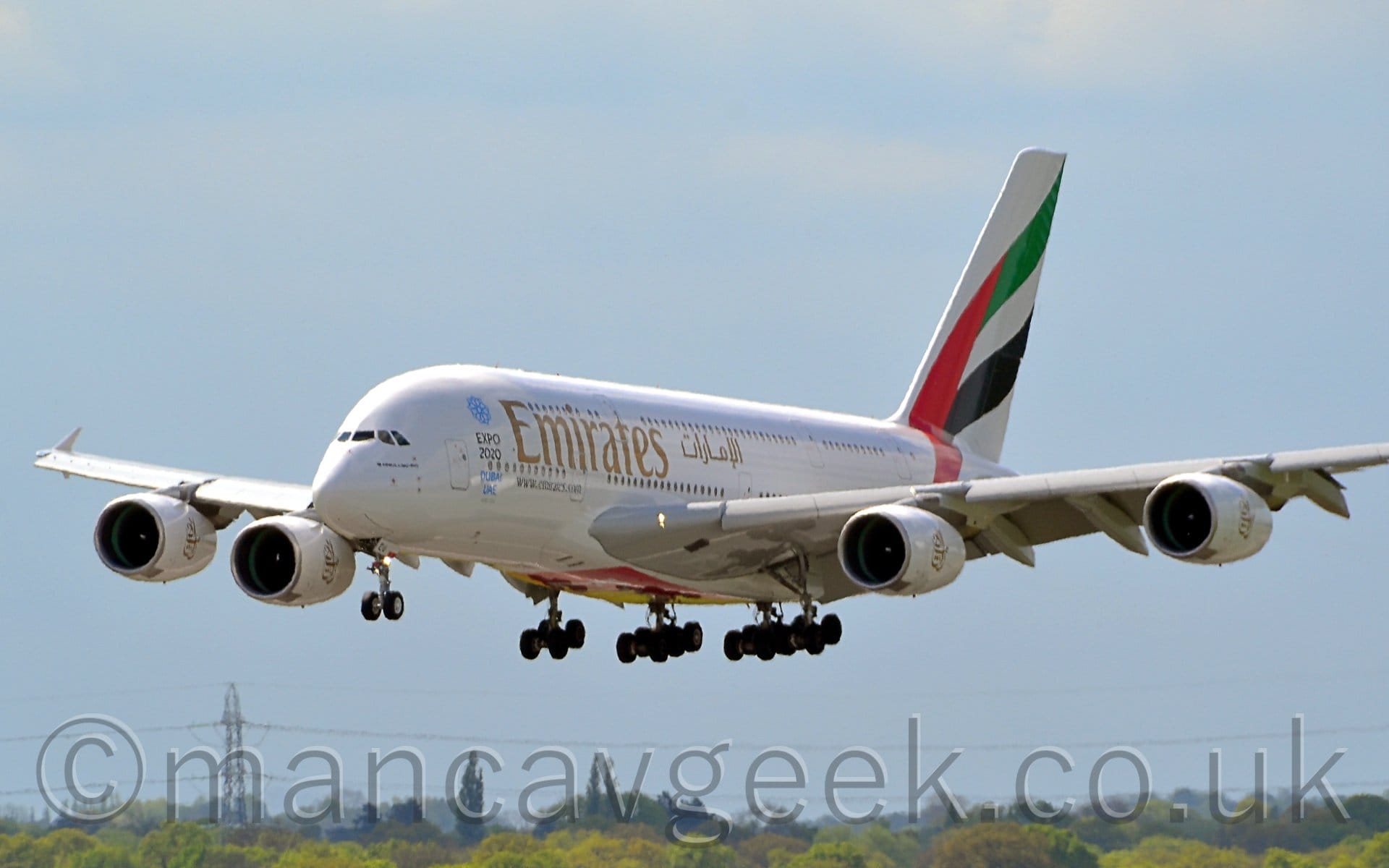 Side view of a very large, white, 4-engined jet airliner flying from right to left at a very low altitude, with undercarriage lowered and flaps extended from the rear of the wings, suggesting it is about to land. The plane is mostly white, with large golden "Emirates" titles  in English on the upper forward fuselage, just in front of smaller titles in an Arabic script. There are smaller, black, "Expo2020" titles slightly forward of the main logo, below the blue outline of a flower and above "blue "Dubai UAE" titles. The 4 white engine pods under the wings hacve large golden logos that are essentially a re-arranged version of the Arabic version of the "Emirates" titles. The planes tail is covered in a large, seemingly waving United Arab Emirates flag (a vertical red bar, with green, white, and black horizontal stripes). Trees and bushes in the distance line the bottom of the frame, under hazy grey skies.