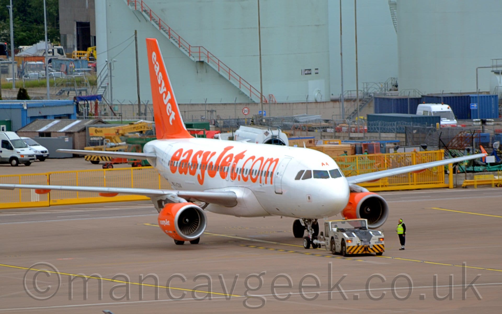 Side view of a white, twin engined jet airliner facing to the right but being pushed back to the left by a white tug. The plane has large, orange, billboard-style "EasyJet[dot]com" titles on the fuselage, with similar white titles on the orange engine pods, and diagonal white titles on the orange tail. The orange letters "AA" are above the centre of the cockpit windows. A man wearing a hi-viz jacket  and hi-viz ear protectors is standing next to the tug, in front on one of the planes engines. A yellow metal fence stretches across the background, with an array of airport vehicles behind it, and a large duck-egg blue structure looming over everything at the rear.