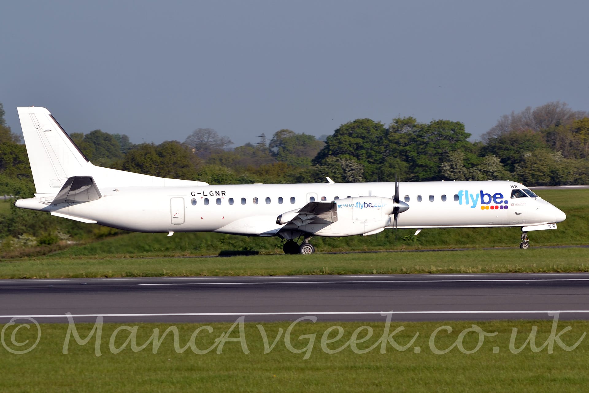 Side view of a white, twin propellor-engined airliner taxiing from left to right off the runway. The plane is almost entirely white, with blue "FlyBe" titles above a series of yellow, red, and purple dots on the forward fuselage, just behind the cockpit. There is the web address "www[dot]flybe[dot]com" in blue on the sides of the engine cowlings. The registration "G-LGNR" is on the upper rear fuselage, in black. The last 2 letters "NR" are on the front of the nose wheel doors, and above the cockpit corner windows, also in black. Green grass lines either side of the black runway in the foreground, with the taxiway this plane is on hidden by grass. More grass in the background leads up to trees in the distance, under blue-grey sky.