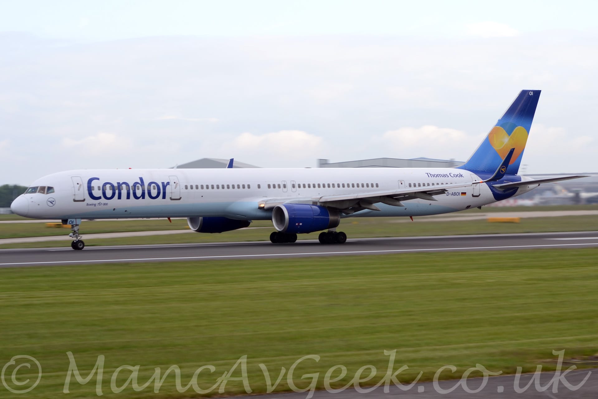 Side view of a white twin engined jet airliner moving from right to left at high speed along a black runway. The plane is mostly white, with a pale blue belly, and dark blue "Condor" titles on the forward fuselage. The tail is dark blue, dipping down slightly to covr part of the rear fuselage, with a lighter blue section through the middle, containing an orange and yellow love heart logo. There are additional blue "Thomas Cook" titles on the upper rear fuselage, just ahead of the tail. The engine pods are the same dark blue, as are the up-turned wingtips, which have the outline of a yellow circle containing a stylised yellow flying bird. Green grass fills the foreground, with more grass in the background leading up to a pair of grey hangars in the distance, under hazy grey sky.