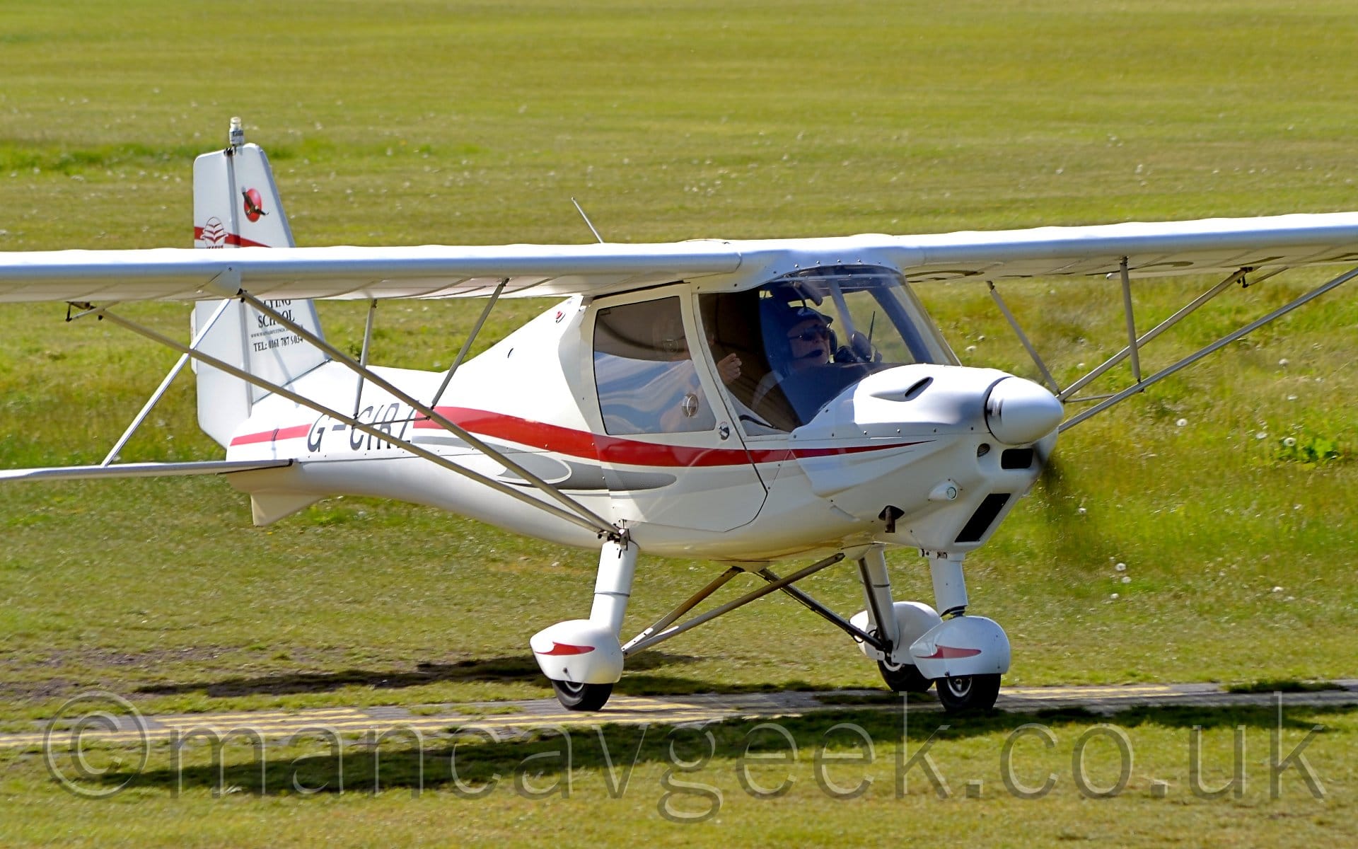 Side view of a white, high-winged, single-engined light aircraft taxiing from left to right on grass. The plane has a red and grey stripe running along the body, and the black registration "G-CIRZ" on the tapered rear fuselage. 2 men can been seen in the cockpit through the large windows. Green grass fills the rest of the frame.