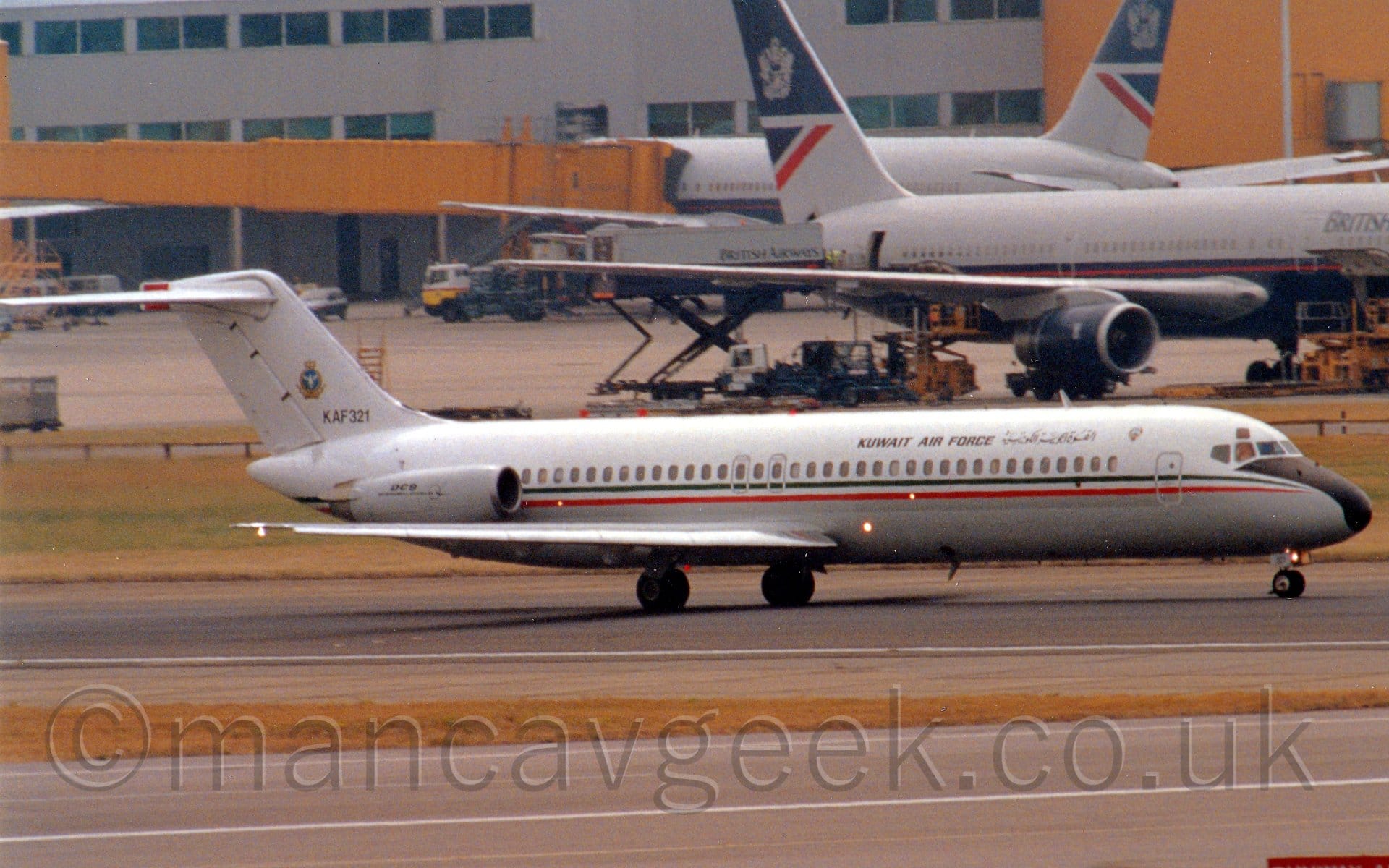 Side view of a twin engined jet airliner moving from left to right along a black runway. The plane has a white top half, a grey lower half, and a thin green, white, and red stripe running along the body. There are black "Kuwait Air Force" titles on the upper forward fuselage, in an Arabic script at the front, and in English towards the middle. The serial "KAF321" is on the lower part of the tail, under a gold, blue, and white logo. A taxiway is at the bottom of the frame, separated from the runway by a wedge of brown grass, with a grey building with yellow air bridges stretches across the frame, the air bridges attached to a pair of grey and dark blue jet airliners .