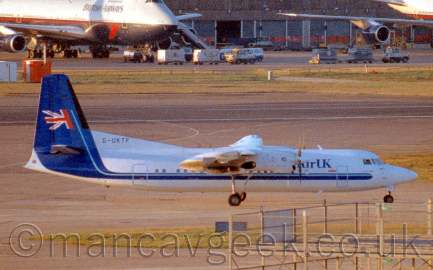 Side view of a white, high-winged, twin propellor-engined airliner taxiing from left to right. The plane is mostly white, with a thin blue stripe running along the body just below the passenger cabin windows, sweeping up the fill the tail. There are blue "Air UK" titles on the upper forward fuselage, and half of a UK flag waving on the tail. The black registration "G-UKTF" is on a white fillet at the front of the base of the tail. Grey concrete apron fills the foreground, with more concrete and grass leading up to a grey building in the distance, with a very large, grey and dark blue 4 engined jet airliner parked on the top left of the frame.