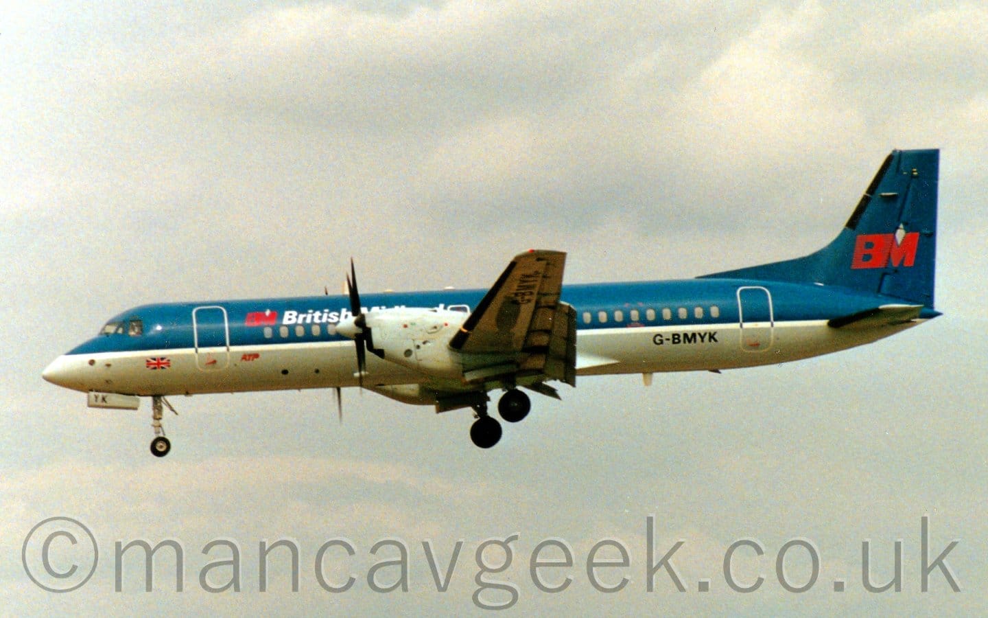 Side view of a blue and grey, twin propellor-engined airliner flying from right to left at a very low altitude, with undercarriage lowered and flaps extended from the rear of the wings, suggesting it is about to land. The plane has dark blue upper surfaces and a grey belly, with white piping, and white "British Midland" titles on the upper forward fuselage next to a red "BM" logo. This logo also appears in larger form on the tail. The registration "G-BMYK" is on the lower rear fuselage in black. Grey sky fills the rest of the frame.