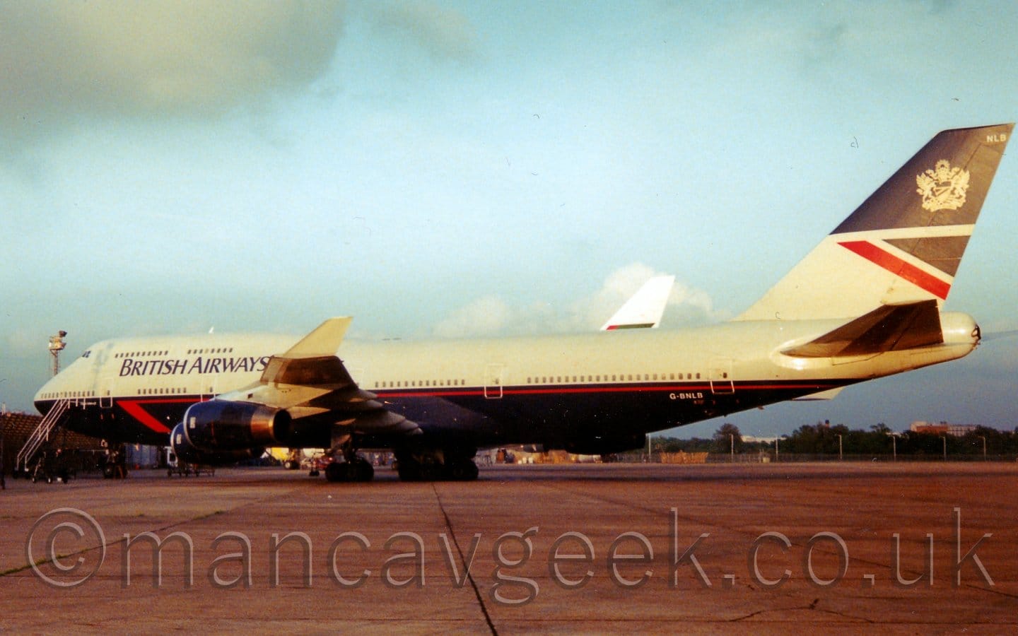Side view of a grey and dark blue, very large, 4 engined jet airliner parked facing to the left. The top half of thew plane is grey, with a dark bluye lower half, with some red piping on the blue section. There are black "British Airways" titles on the upper forward fuselage. The tail is dark blue on the top half, the bottom being grey with a red and dark blue triangle under the blue top half. Grey concrete apron fills the foreground, running off into the distance, eventually giving way to trees and buildings. The white tail of another airliner can be seen above the rear fuselage of this plane. Pale blue sky fills the rest of the frame, with fluffs of grey cloud on the left.