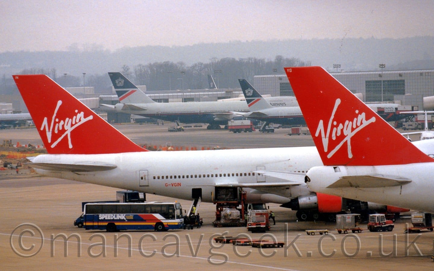 Close-up of the rear fuselage and tails of a pair of very large, white, 4 engined jet airliners parked facing to the right. The planes are both mostly white, with red tails with white "Virgin" titles, and red engine pods. The further plane has the red registration "G-VGIN" on the lower rear fuselage. Baggage trucks, coaches, and other airport vehicles are scattered around the apron around them. A grey terminal building stretches across the background, with grey and dark blue airliners parked in front. Trees slowly vanish into haze in the distance, under grim grey skies.