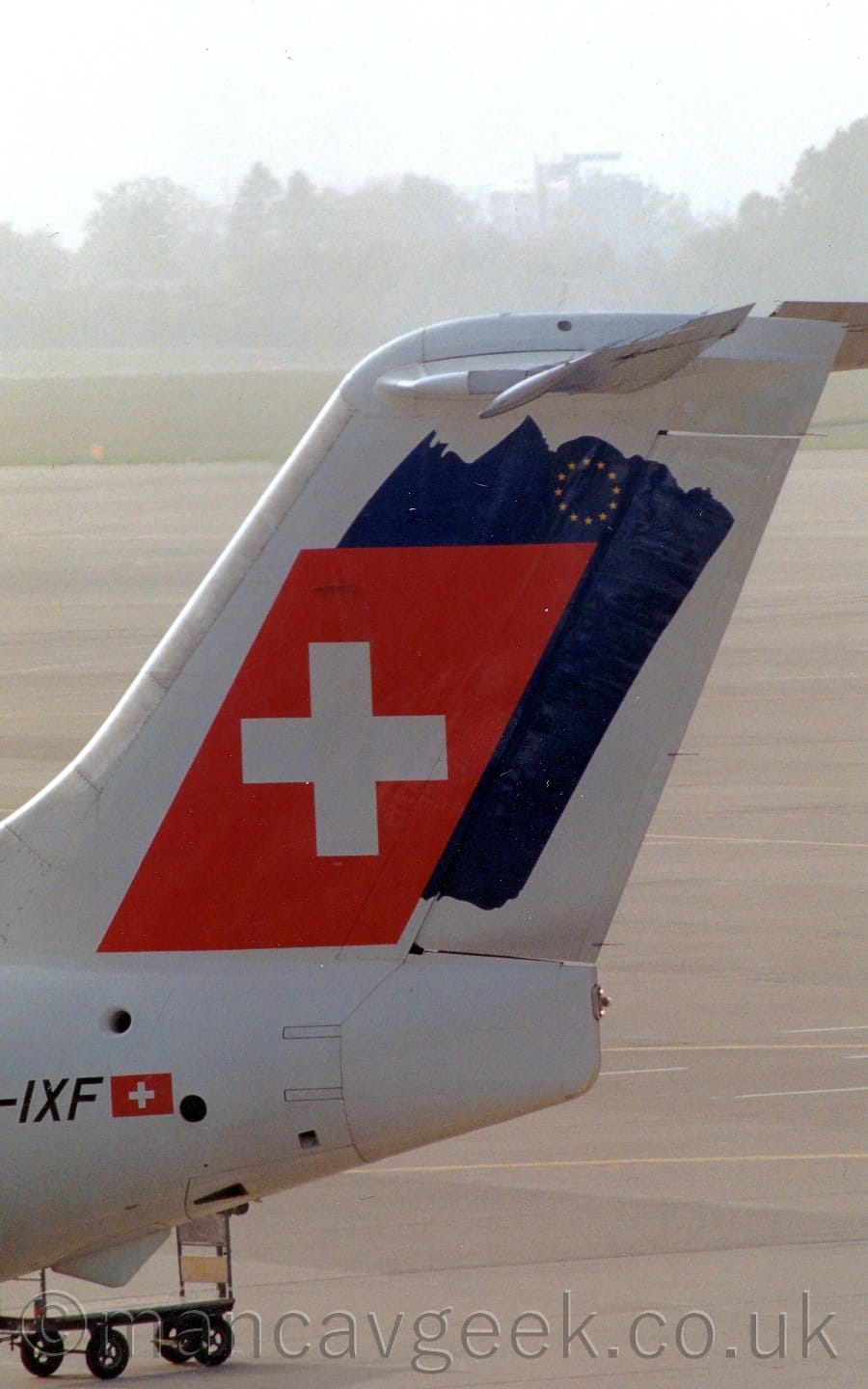 Close-up of the tail of a jet airliner parked facing to the left. The plane is mostly white, with a red, angular rectangle with a white cross in the middle, over an uneven patch of dark blue paint with a small circle of gold stars at the top, covering most of the tail. The black partial registration "-IXF" is on the rear fuselage, next to a small Swiss flag. Grey concrete apron fills most of the rest of the frame, with trees at the top vanishing into haze in the distance.