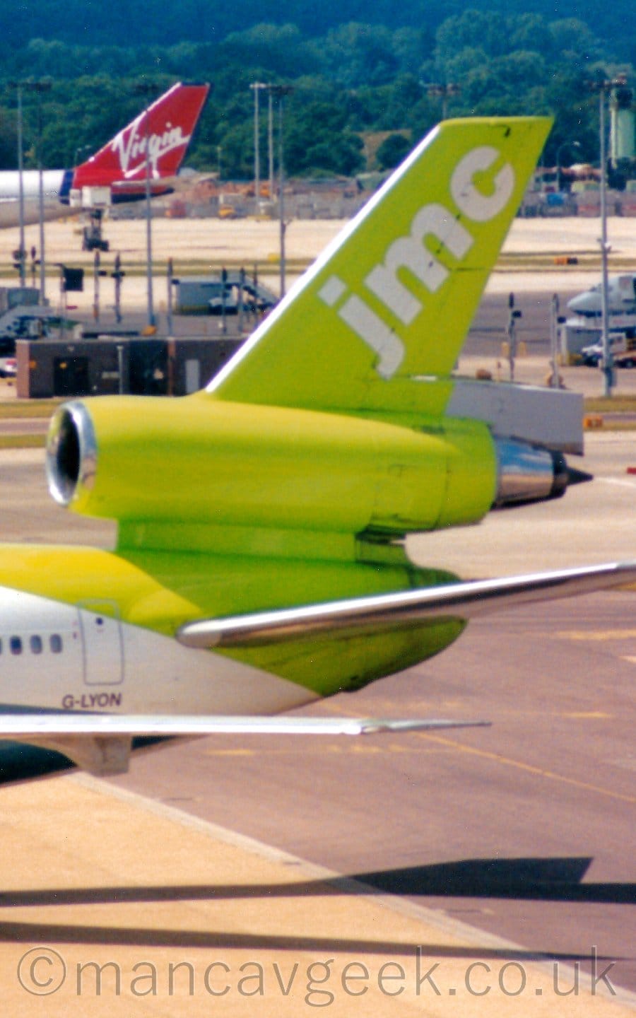 Close-up of the tail and centre engine of a 3 engined jet airliner taxiing from right to left. The plane's fuselage is white, with a bright, lime green tail with diagonal, white "JMC" titles on the top section. The registration "G-LYON" is on the lower rear fuselage in black. Tall lighting stands are dotted around the background, with the red and white tail of another jet aircraft at the top left.