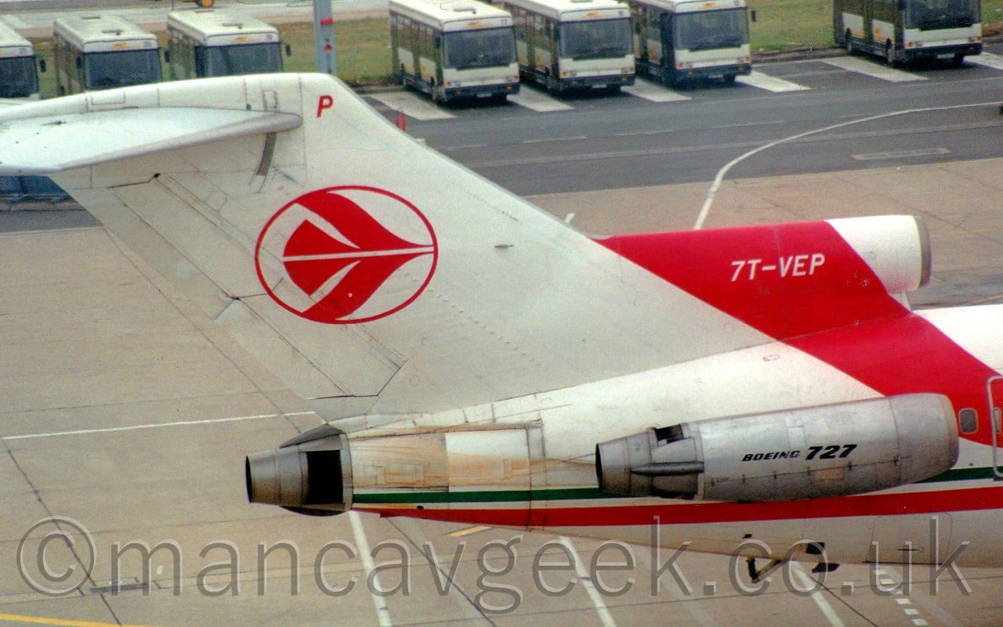 Close-up of the white tail of a 3-engined jet airliner with the engines mounted on the rear fuselage, taxiing from left to right. There is a thin red and green stripe running along the body, with a wider, red diagonal stripe coming forward from the top of the centre-mounted engine intake, with the white registration "7T-VEP". The tail has the red outline of a circle with a stylised red flying bird in the middle, and a red letter "P" right at the top. The side-mounted engine pods are grey, with black "Boeing 727" text on the sides. Grey concrete apron fills the background, with a black roadway tacked on to the far side, with blue buses parked side by side along the far edge.