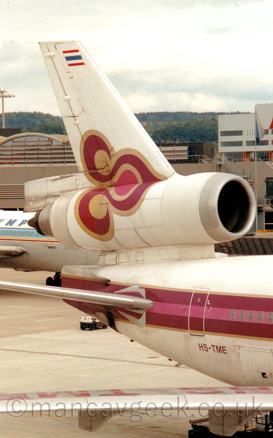Close-up of the rear fuselage and tail of a 3 engined jet airliner parked facing to the right. The plane is mostly white, with red and pink stripe outlined in gold running along the body, along with a red and pink flower design design also outlined in gold. The ed registration "HS-TME" is on the lower rear fuselage. A brown terminal building stretchews across the frame in the background, with trees and rolling hills in the distance, under cloudy grey sky.