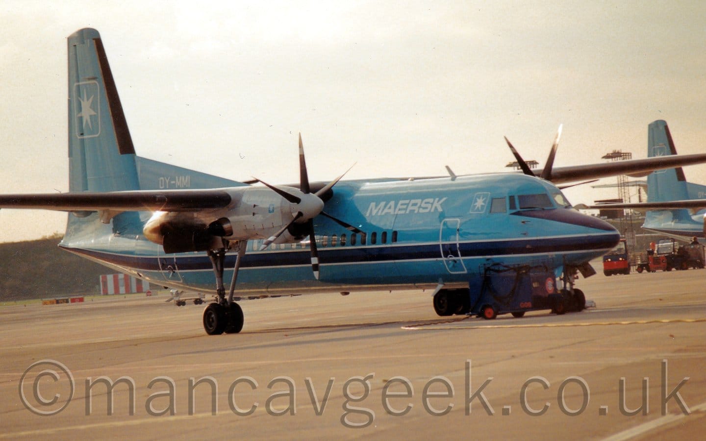 Side view of a high-winged, twin propellor-engined airliner parked facing to the right. The plane is a light blue, with a dark blue stripe outlined in white running along the body under the passenger cabin windows, and white "Maersk" titles on the upper forward fuselage.The white registration "OY-MMI" is on a fillet at the front of the tail. There is the white outline of a square containing a 7-pointed star on the tail, repeated in smaller form just aft of the cockpit windows.grey concrete apron fills the foreground, with a grass berm in the background coming in from the left.The tail of a similar plane can be seen on the right of the frame. Bright but hazy skies fill the rest of the frame.