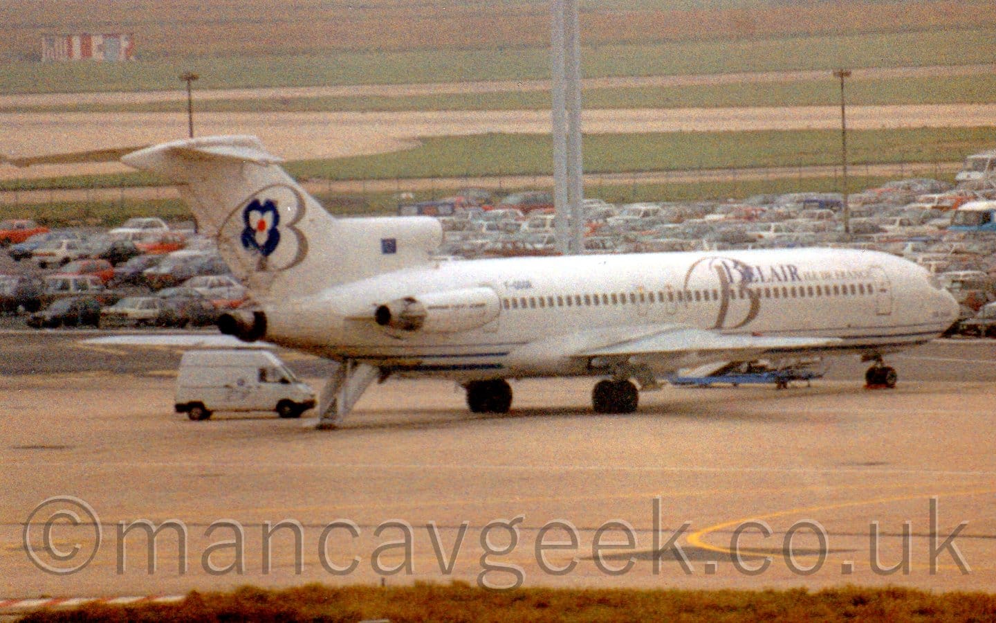 Rear shot of a 3 engined jet airliner with the engines mounted on the rear fuselage. There are blue "BelAir" titles on the upper forward fuselage, overlaid with a large grey letter "B". A similar letter "B" is on the tail, next to a dark blue, white, and red stylised flower. The black registration "F-GGGR" is on the upper rear fuselage, under the tail. A set of built-in airstairs is coming out of the rear fuselage, under the tail, next to a white van. Grey concrete apron fills the foreground, with parked cars and green grass leading off into the distance in the background.