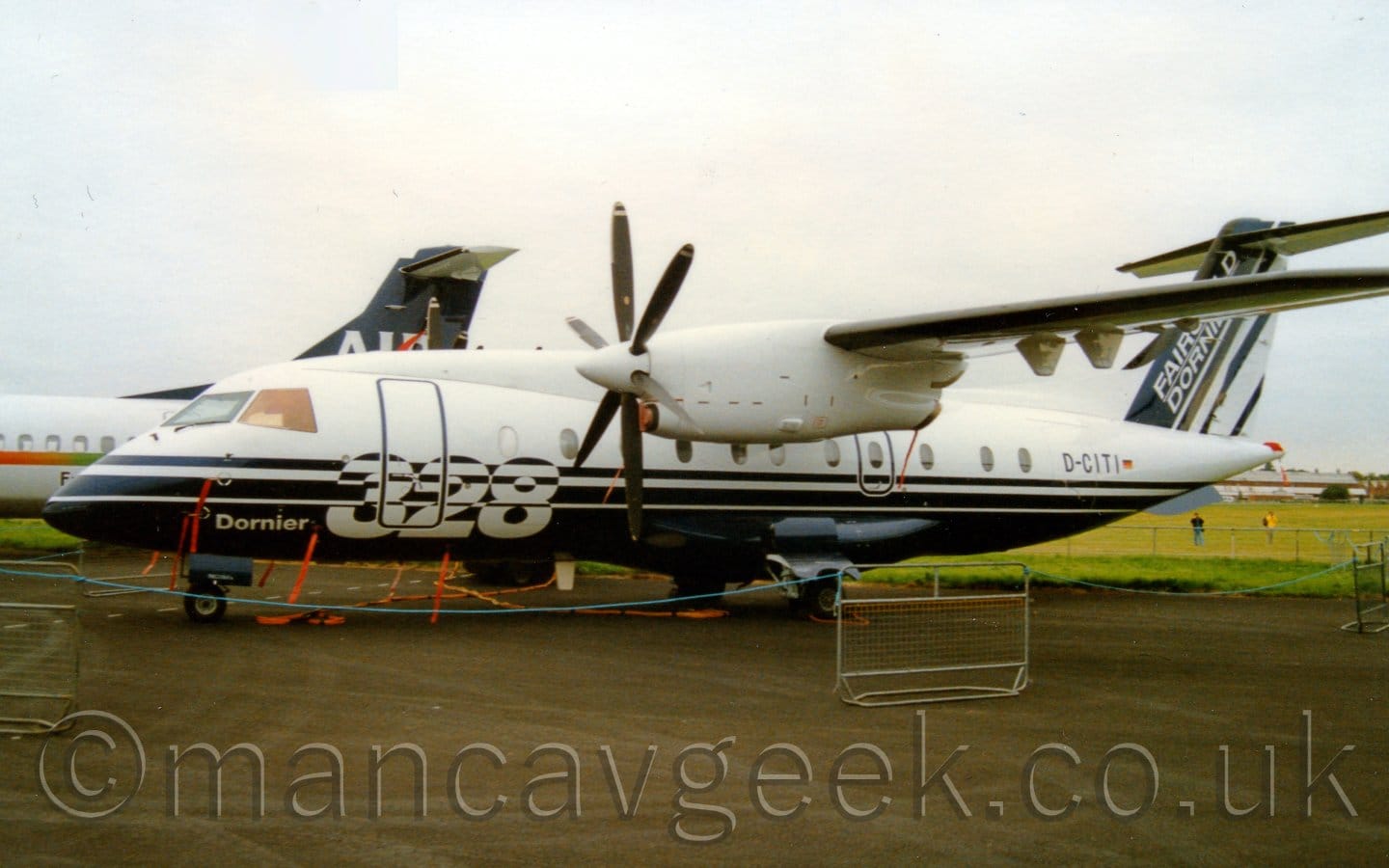 Side view of a high-winged, twin propellor-engined airliner parked on black tarmac facing to the left. The plane has white upper surfaces, with dark blue lower surfaces, and a large white number "328" on the lower forward fuselage. The black registration D-CITI is on the upper rear fuselage, under the dark blue and white tail, which has white "Fairchild Dornier" at the front. Sections of metal fence joined with long pieces of blue rope are placed in front of the plane. The rear fuselage and tail of another airliner can be seen on the left, with grey sky filling the rest of the frame.