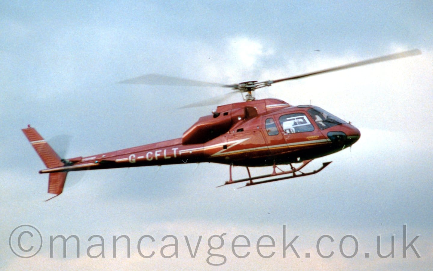 Side view of a brown helicopter with skids for undercarriage flying from left to right at a low altitude. There is a silver stripe running along the body, zig-zagging it's way up to the tailboom, where it is overlaid with the silver registration "G-CFLT". Patchy white clouds with hints of blue fill the rest of the frame.