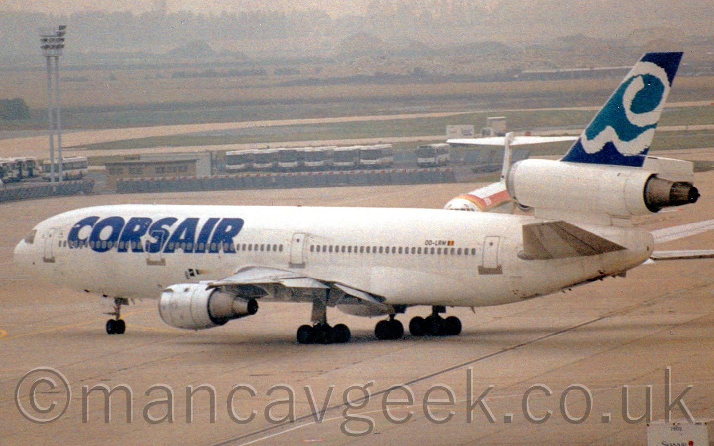 Side view of a 3 engined jet airliner taxiing from right to left. The plane is mostly white, with large, blue, billboard-style "Corsair" titles on the forward fuselage, and blue, aquamarine, and white stylised waves on the tail. Grey concrete apron fills the foreground, with rows of airport buses lined up side-by-side between taxiways in the background. Fields beyond there lead up to trees in the distance, under hazy grey sky.