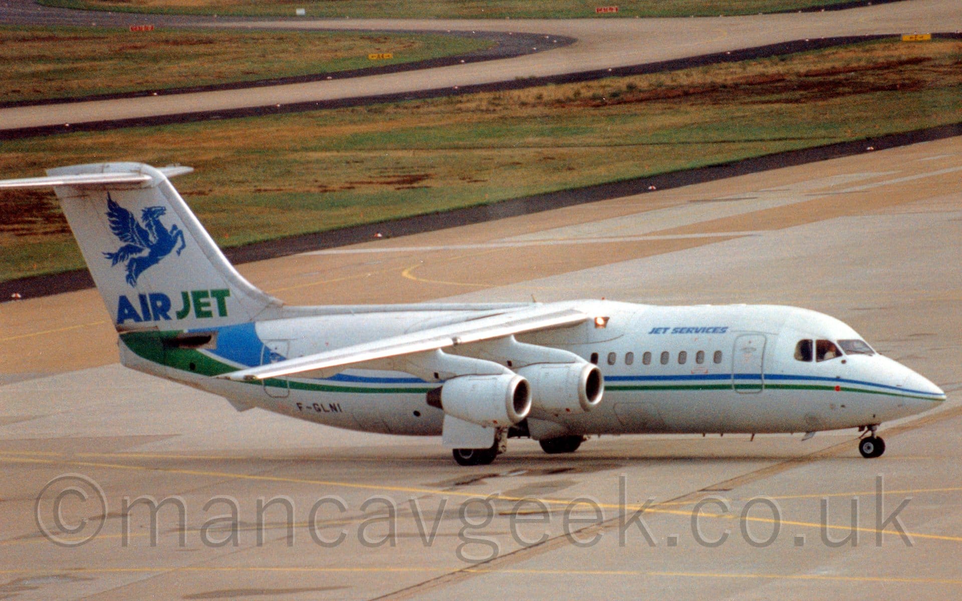 Side view of a high-winged, 4 engined jet airliner taxiing from left to right. The plane is mostly white, with a green and blue stripe running along the body, getting wider and swooping slightly up towards the tail as it moves rearwards. There are small, light blue "Jet Services" titles on the upper forward fuselage, and the black registration "F-GLNI" on the lower rear. There are blue and green "AirJet" titles at the base of the tail, under a large, blue Pegasus. Grey concrete apron fills most of the foreground,, with more apron in the background, with large grassed areas lining the sides of several taxiways.
