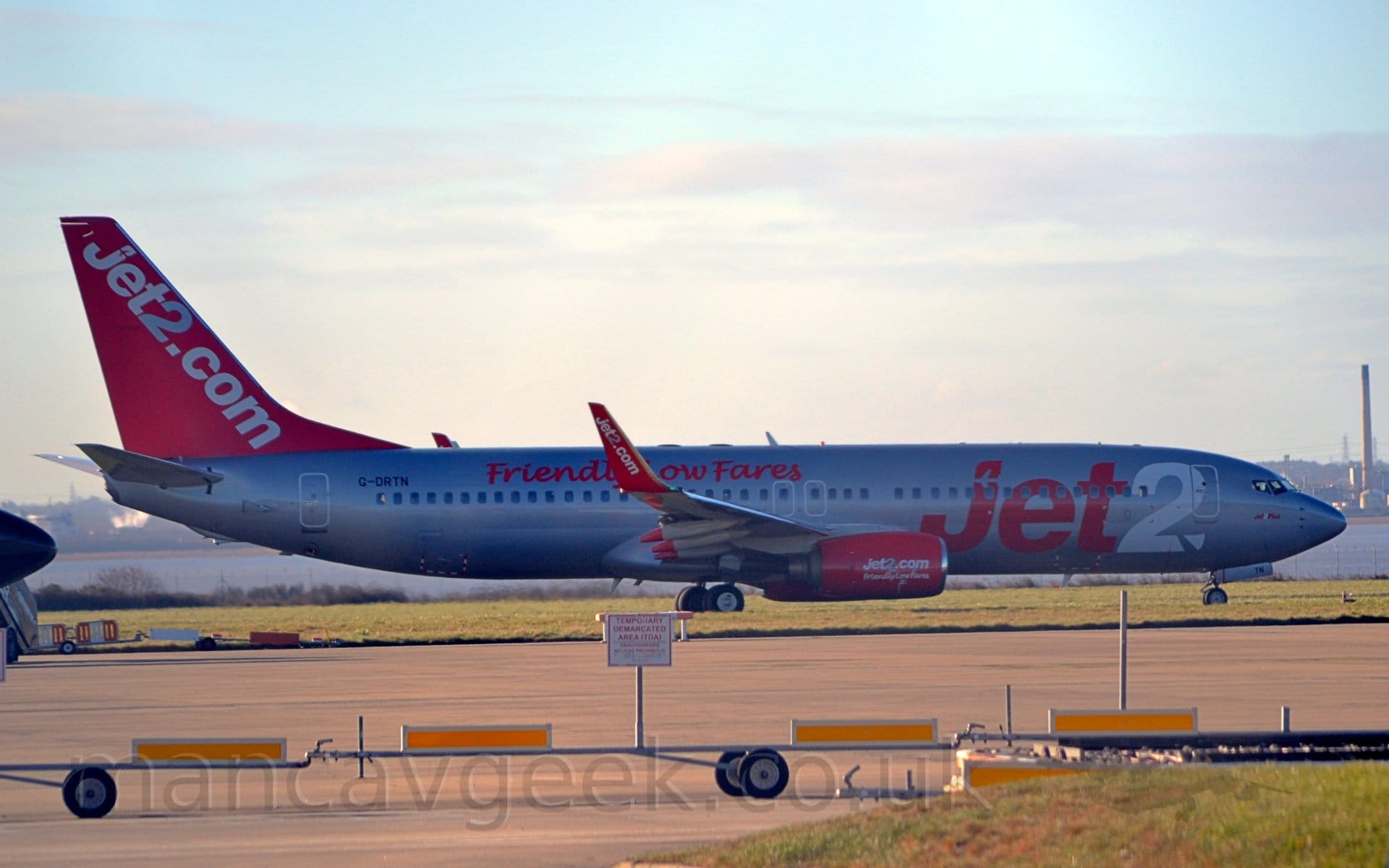 Side view of a twin engined jet airliner taxiing from left to right. The plane is an almost silvery-grey, with large "Jet2" titles on the forward fuselage, the number "2" being white. There is large red, seemingly hand-written "Friendly Low Fares" text on the upper rear fuselage, just forward of the black registration "G-DRTN". The tail is red, with diagonal white "Jet2.com", with the number "2" in grey, a design repeated on the red upturned wingtips. The engine pods mounted under the wings more "Jet2.com" titles, anlong with white "Friendly Low Fare" text. Grey concrete apron fills the foreground, with a river stretching across the frame in the background, with an industrial landscape on the far side, under bright but hazy skies.
