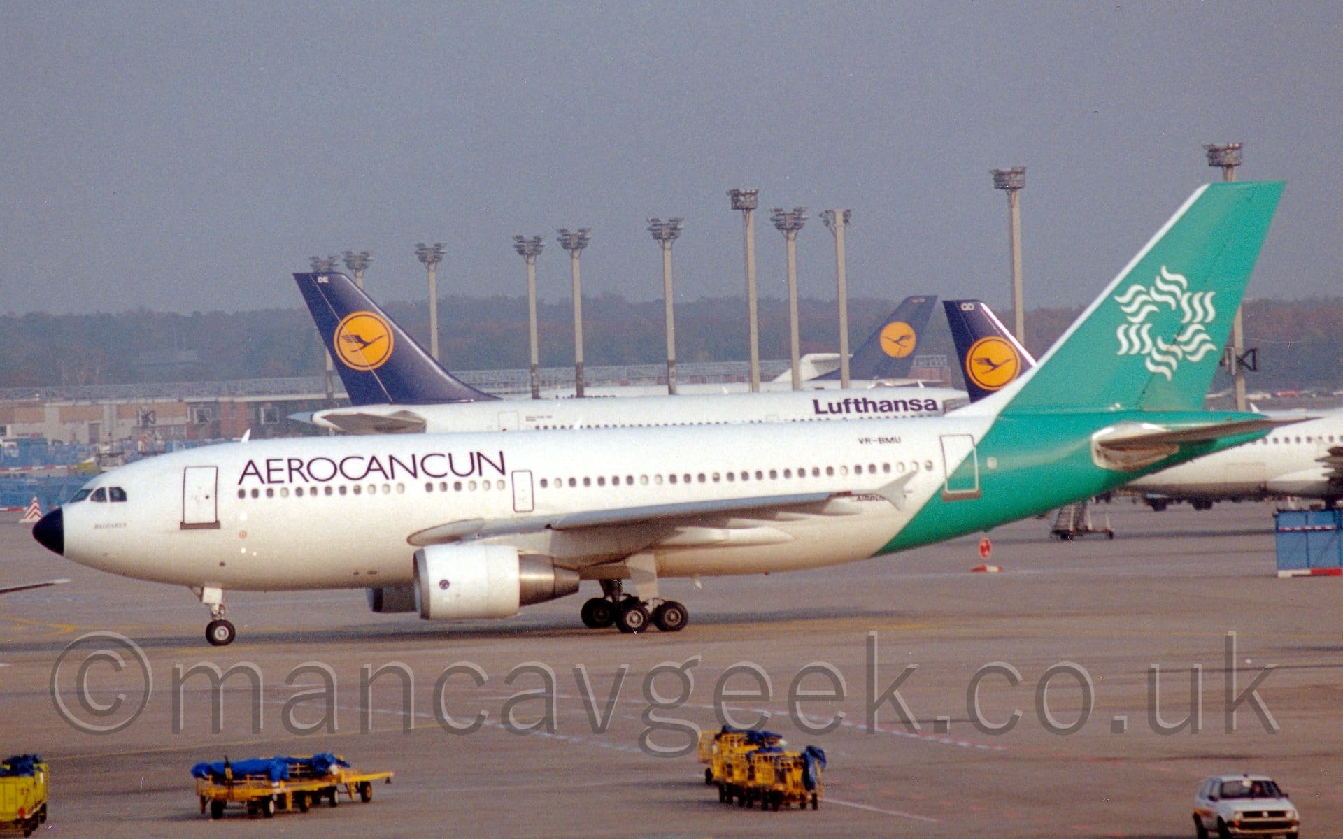 Side view of a twin engined jet airliner taxiing from right to left. The plane is mostly white, with black "AeroCancun" titles on the upper forward fuselage. The rear fuselage are a minty green, with a white stylised flower logo made up of six sets of 3 wavy lines arranged in a circle. Grey concrete apron fills the foreground, with yellow luggage carts with blue covers parked at the side of a runway marked on the concrete. In the background, several white jet airliners with blue and yellow tails are parked amidst some tall lighting towers, with trees in the distance, under grey sky.