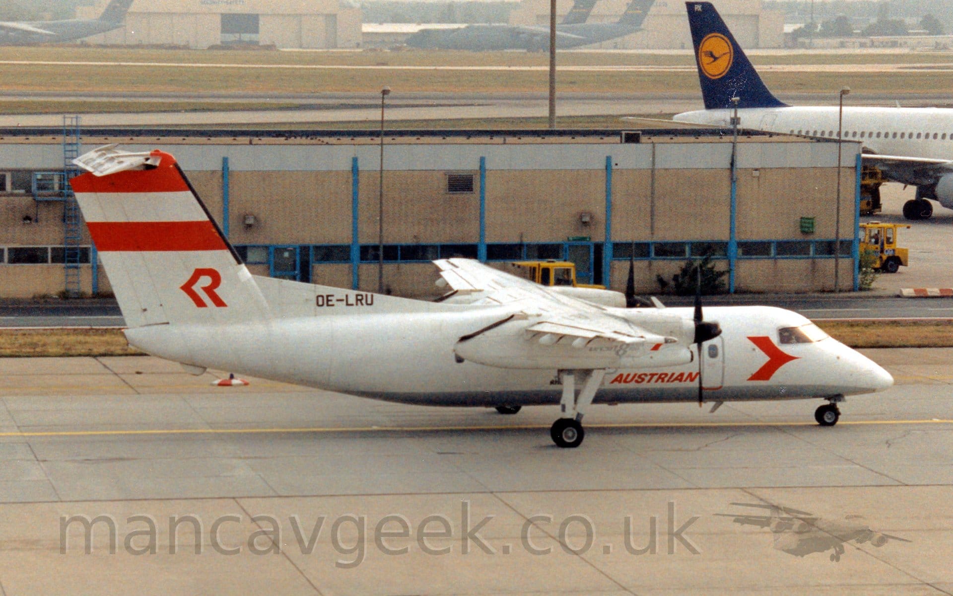 Side view of a high-winged, twin propellor-engined airliner taxiing from left to right. The plane is mostly white, with a grey belly, and red "Austrian" titles on the lower forward fuselage, with a red arrowhead just below and aft of the cockpit. There are a pair of thick red stripes at the top of the white tail, with a stylised red upper-case letter "R" at the base. A pale brown building stretches across the background, with a white jet airliner with a blue and yellow tail just taxiing out of frame at the top right. Large, green, 4 engined military cargo planes are clustered around a cream coloured hangar in the distance, slowly vanishing in to haze.