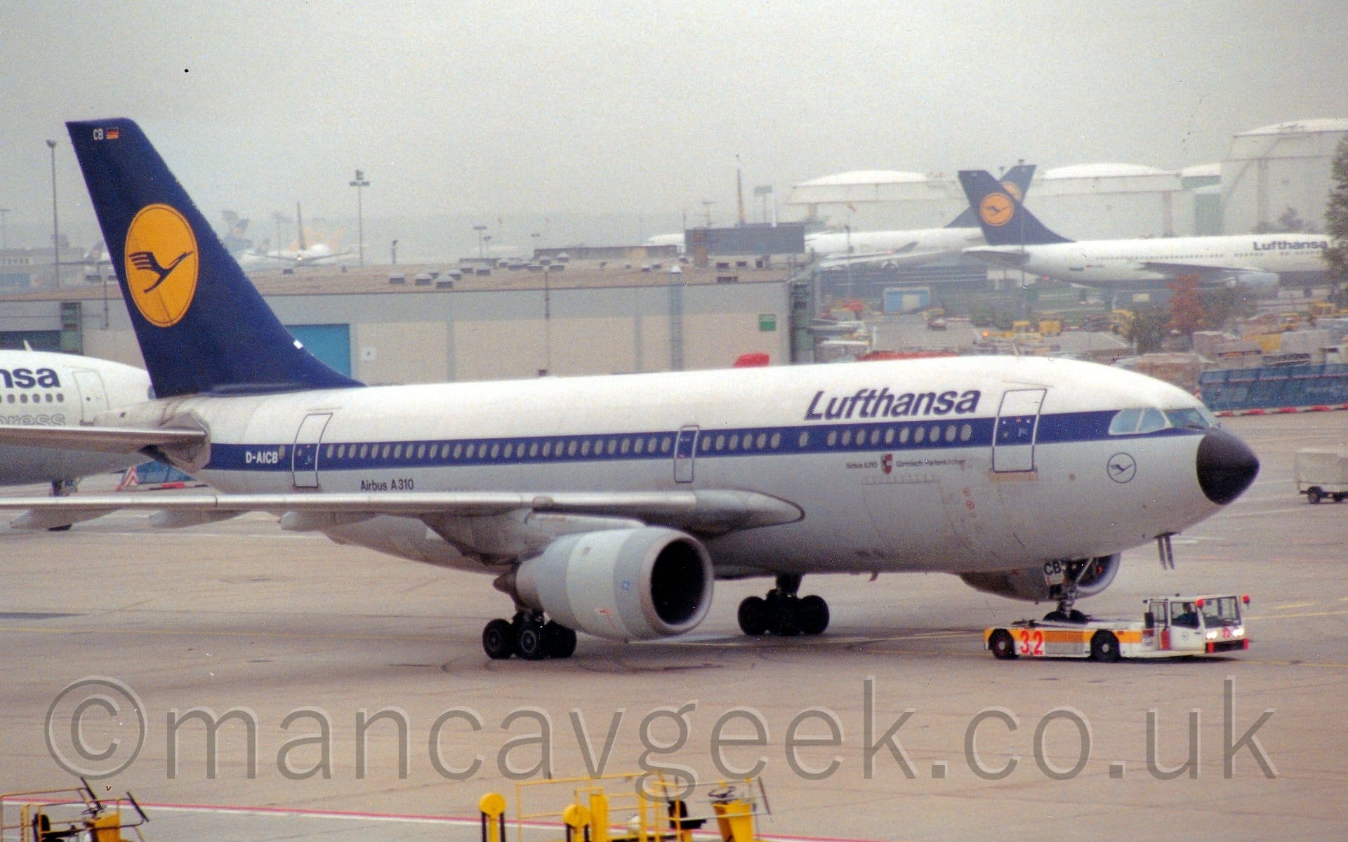 Side view of a twin engined jet airliner being towed from left to right by a low-slung white tug with a yellow stripe along the body, which has clasped and raised the plane's nosewheel in order to move it around. The plane has a white upper body, with a grey belly, and a dark blue stripe running covering the passenger cabin windows. There are dark blue "Lufthansa" titles on the upper forward fuselage, with the dark blue outline of a circle containing a stylised blue bird under the cockpit windows. The white registration "D-AICB" is on the blue stripe at the rear of the fuselage, with black text "Airbus A310" on the lower rear body. The tail is dark blue, with a yellow circle containing a stylised blue flying bird, similar to the logo under the cockpit. Grey concrete apron fills the foreground, with grey buildings and white planes with blue and yellow tails filling the background under grey sky.