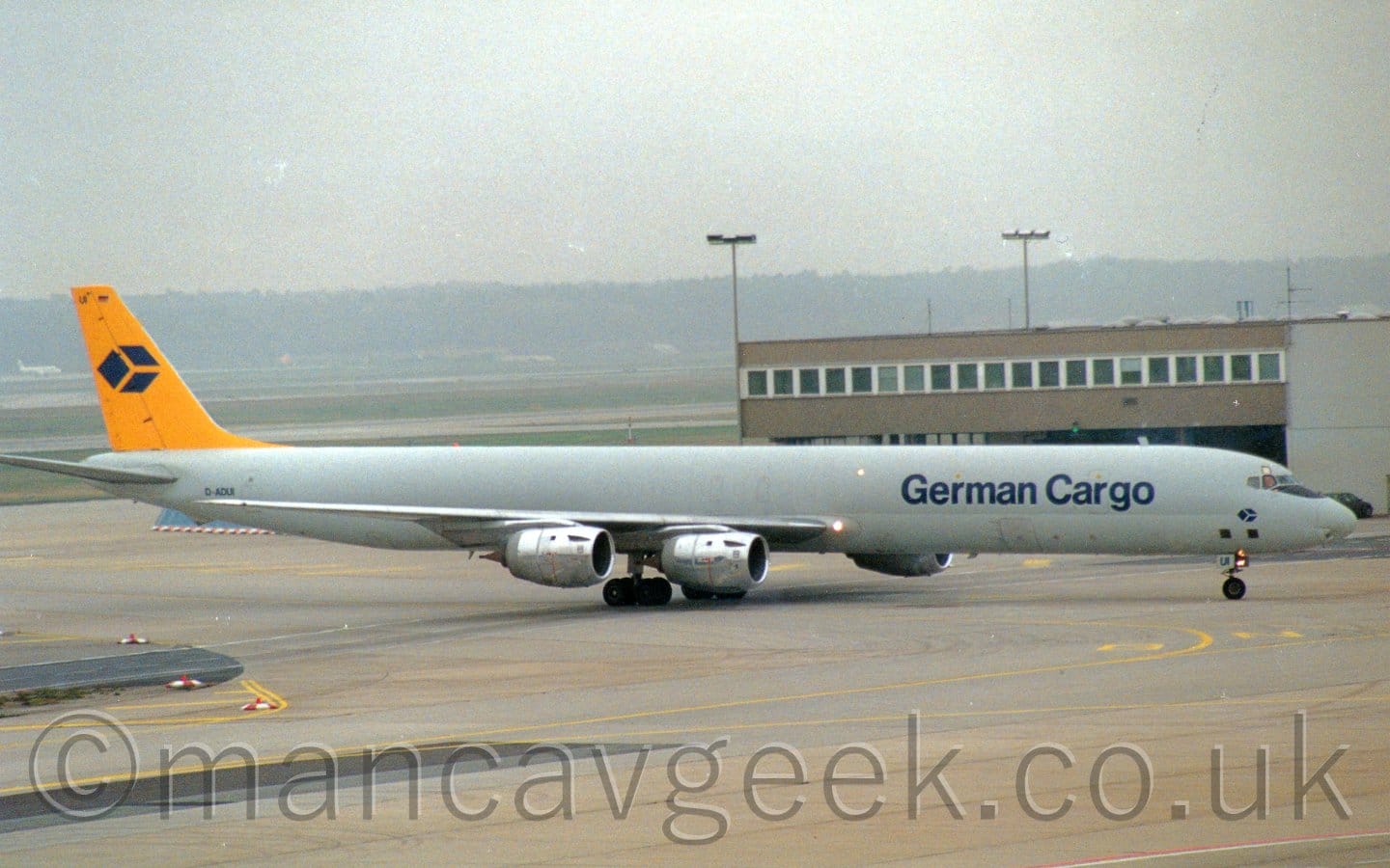 Side view of a long, thin, 4 engined jet airliner taxiing from left to right. The plane is mostly a dark grey, with a lighter grey belly, and dark blue "German Cargo" titles on the forward fuselage. The tail is bright yellow, with a logo of a 3-D blue box in the middle - the same logo appears in a smaller form under the cockpit windows. Grey concrete apron fills the foreground, with a brown building stretching half way across the frame from the right in the background. On the left, grass and runways lead up to trees in the distance, under hazy grey skies.
