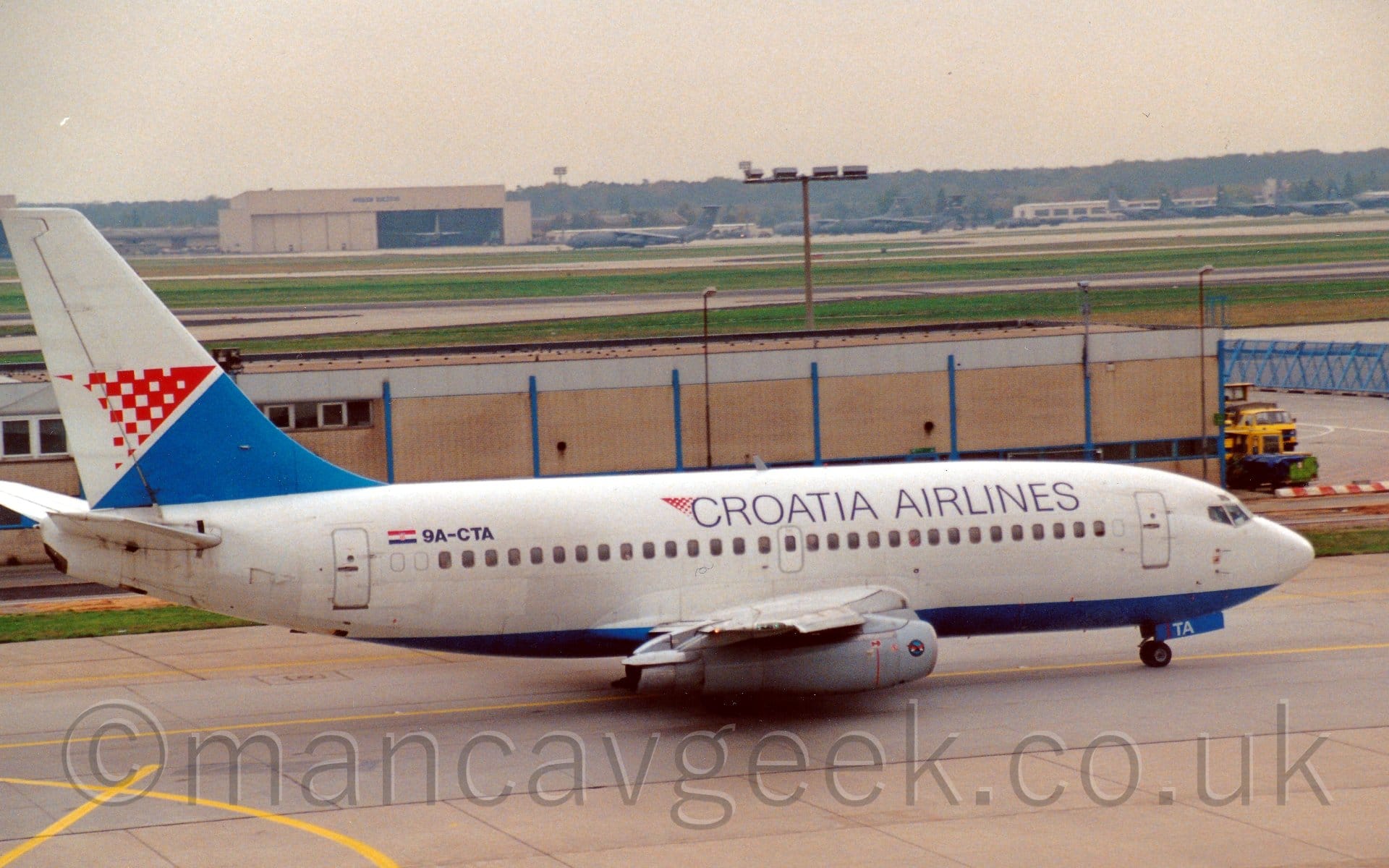 Side view of a twin engined jet airliner taxiing from left to right. The plane is mostly white, with a pale blue belly. There are black "Croatia Airlines" titles on the upper forward fuselage, with the black registration "9A-CTA" on the upper rear, next to a small Croatian flag. The tail is mostly white, with a blue triangle at the base, below a slightly smaller red and white triangle. A low, brown building stretches across the frame from the left, immediately behind this plane, with large areas of green grass separating black runways and grey taxiways that lead up to cream-coloured hangars in the distance, with large green military transports parked around it. Trees line the horizon, under bright but hazy skies.