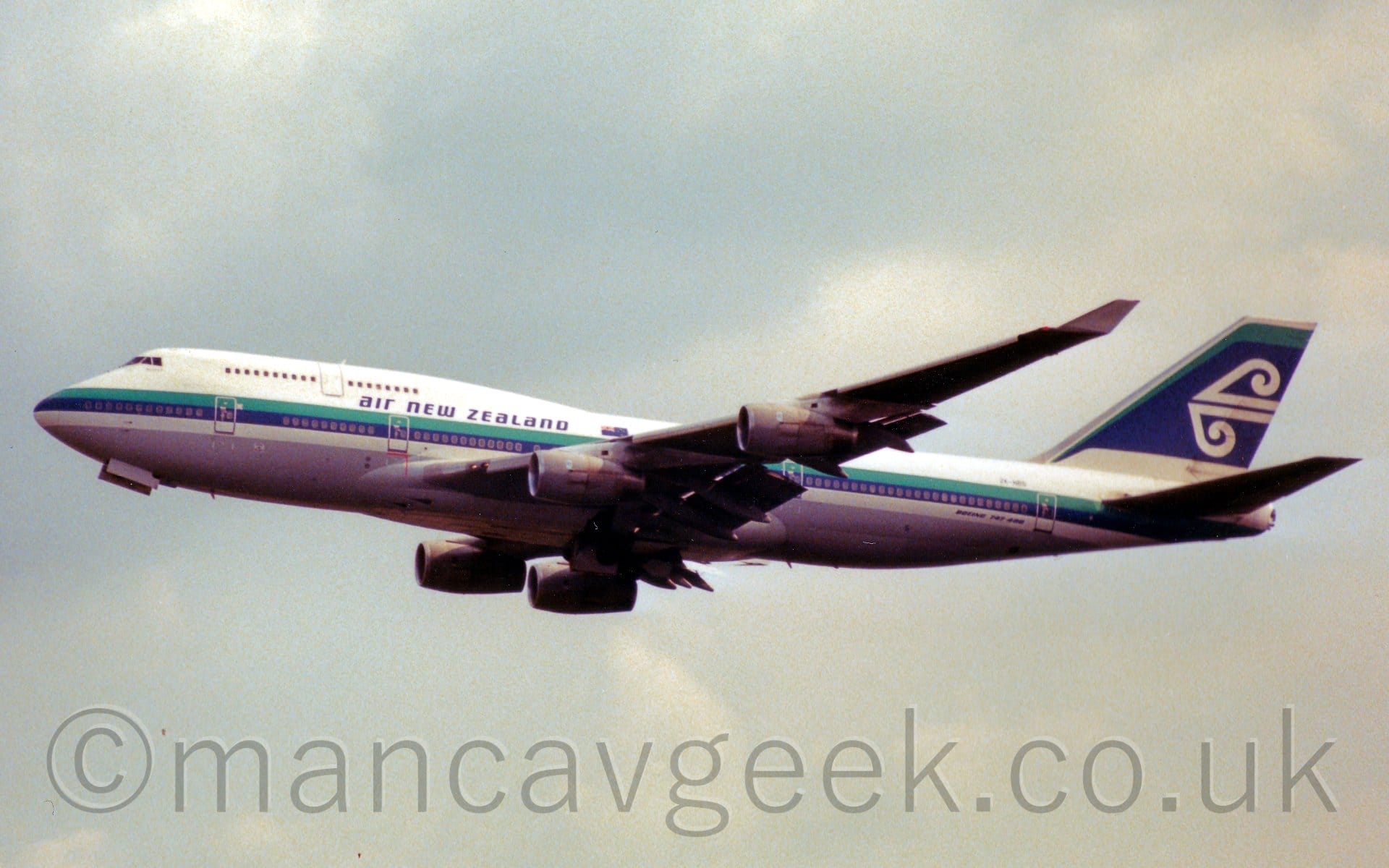 Side view of a very large, 4 engined jet airliner flying from right to left at a low altitude, with flaps extended from the rear of the wings, the undercarriage already tucked away, and the nose raised sharply, suggesting it has just taken off. The plane is mostly white, with a grey belly, and a green and blue stripe running along the body, covering the lower deck cabin windows., with blue "Air New Zealand" titles at the rear of the upper deck. The tail is mostly dark blue, with a green streak at the front and top, and a stylised white wave in the middle. Grey cloudy sky with patches of blue fill the rest of the frame.
