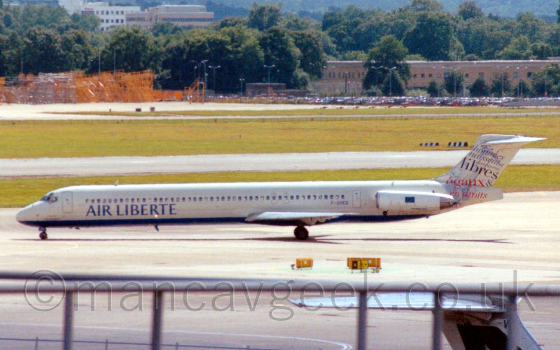 Side view of a twin engined jet airliner with the engines mounted on the sides of the rear fuselage, taxiing from right to left. The plane is mostly white, with a blue belly, and blue "Air Liberte" titles on the lower forward fuselage. The tail is mostly white, with blue and red text, mostly French words mostly related to "Liberty". In the foreground, the top of a railing at the edge of the viewing terraces can be seen at the bottom of the frame, with large areas of grass divided by runways and taxiways in the background, leading up to brown buildings nestled in trees in the distance.