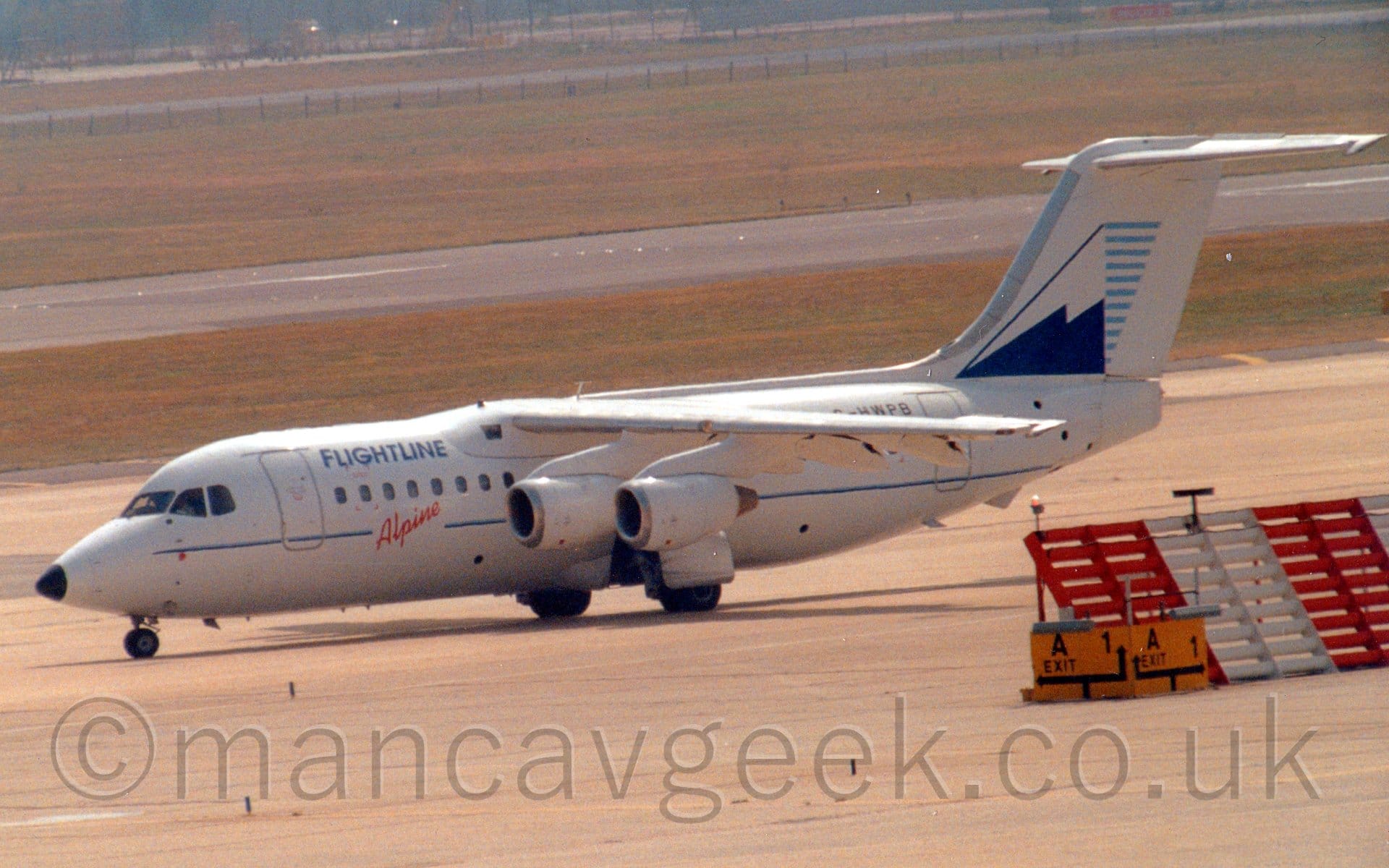 Side view of a high-winged, 4 engined jet airliner taxiing from right to left, with flaps extended from the rear of the wings, suggesting it is going out to a runway for takeoff. The plane is mostly white, with a thin blue stripe running along the body, overlaid with diagonal red "Alpine" titles below the passenger cabin windows just ahead of the wings, and blue "Flightlines" titles on the upper forward fuselage. The black registration "G-HWPB" is on the upper rear fuselage, partially obscured by the wing. The tail is white, with a blue and white stylised image of a snow-capped mountain, with short, light blue lines on it's leeward side. Grey concrete apron fills the foreground, with an angled red and white metal barrier on the right of the frame, while wide strips of green grass line a black runway in the background.