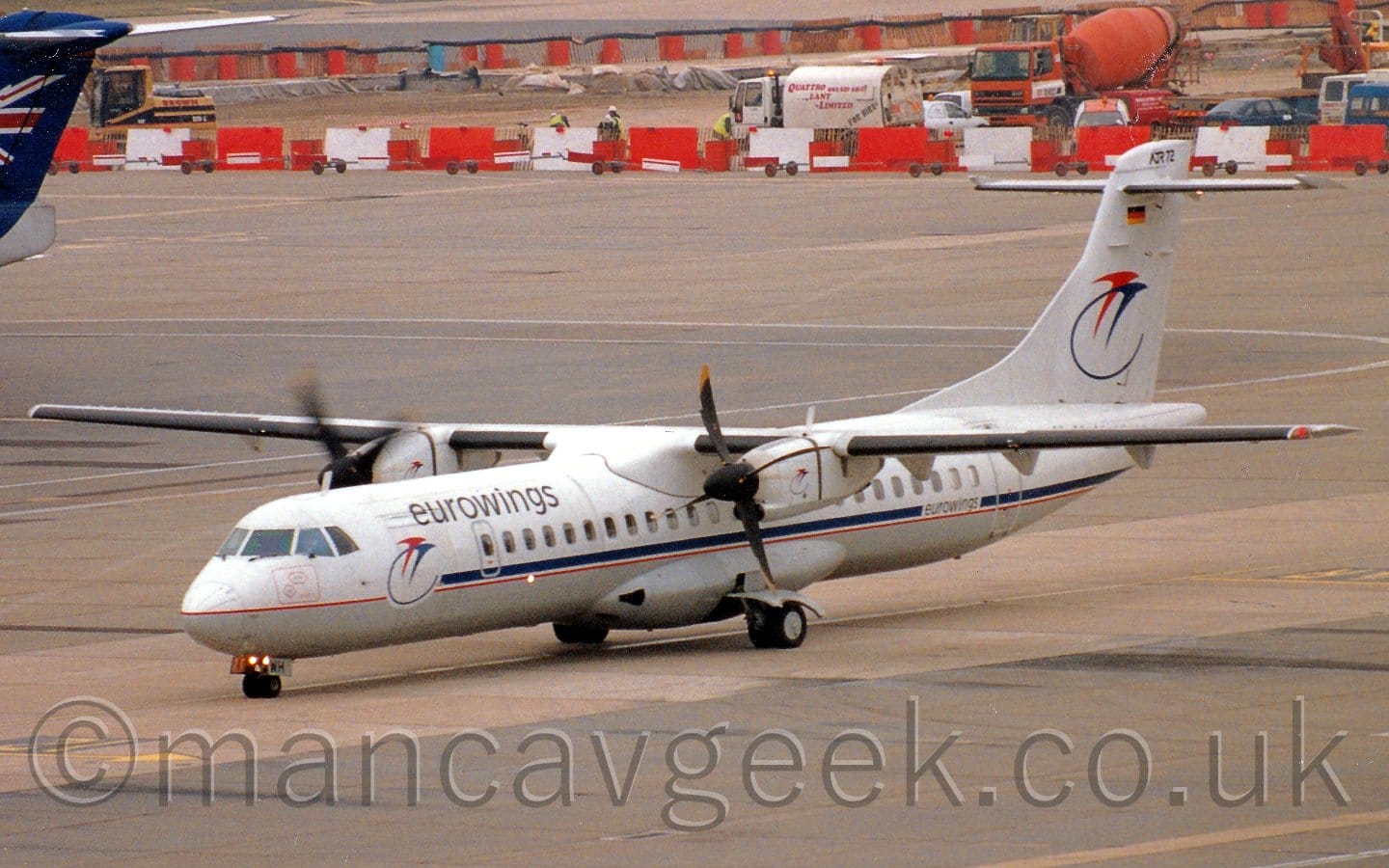 Side view of a high-winged, twin propellor-engined airliner taxiing from right to left. The plane is mostly white, with a blue stripe and red pin-stripe running along the body, as well as a grey belly. There are black "Eurowings" titles on the upper forward fuselage, above a pair of stylised flying bird, one red, one blue with a long tail that forms a semi-circle, a logo repeated on the tail. Grey concrete apron fills the foreground and most of the background, with white and red barriers at the top of the frame denoting a construction zone, with several vehicles inside including an orange concrete truck, and men wearing hi-viz vests and hard-hats.