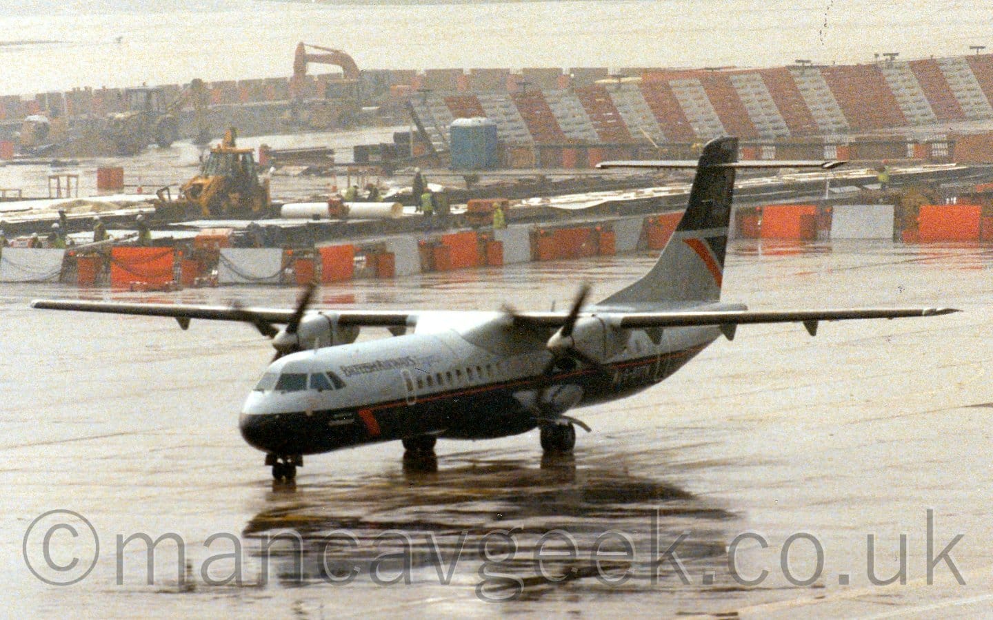 Slightly grainy side view of a high-winged, twin propellor-engined airliner taxiing from right to left. The plane has grey upper surfaces and a dark blue, almost black belly, with black "British Airways Express" titles on the upper forward fuselage. Rain-sodden concrete apron fills the foreground, with a rather damp looking building site outlined with white and red striped barriers in the background.