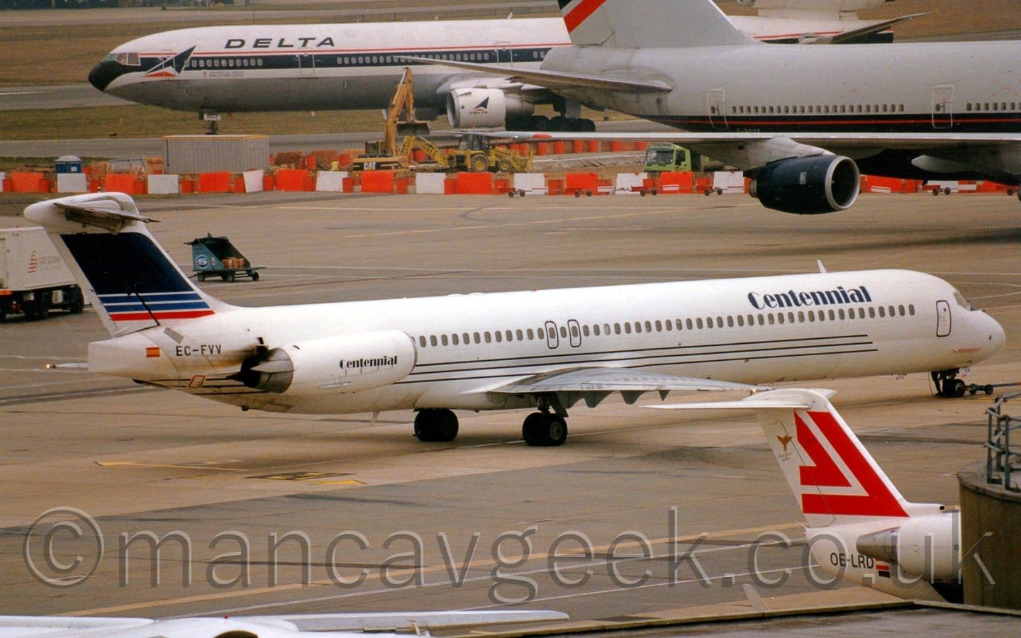 Side view of a twin engined jet airliner with the engines mounted on the sides of the rear fuselage, facing to the right but being pushed back to the left by an out-of-frame tug attached to the planes nosewheel. The plane is mostly white, with a trio of dark blue pinstripes running along the lower fuselage under the passenger cabin windows, and dark blue "Centennial" titles on the upper forward fuselage, repeated on the sides of the engine pods. The black registration "EC-FVV" is on the upper rear fuselage, next to a small image of a Spanish flag, just aft of the engines. The tail is mostly dark blue, with 2 thin lighter blue stripes and a similar red stripe at the bottom, all outlined in white. The foreground is mostly filled with grey concrete apron, with the white and red tail of a small jet airliner poking out from behind the corner of a dark grey terminal engine on the bottom right of the frame. A grey and dark blue, very large 4 engined airliner is taxiing across the frame from left to right in the background, with a white and black 3 engined jet airliner taxiing from right to left towards the runway beyond that.