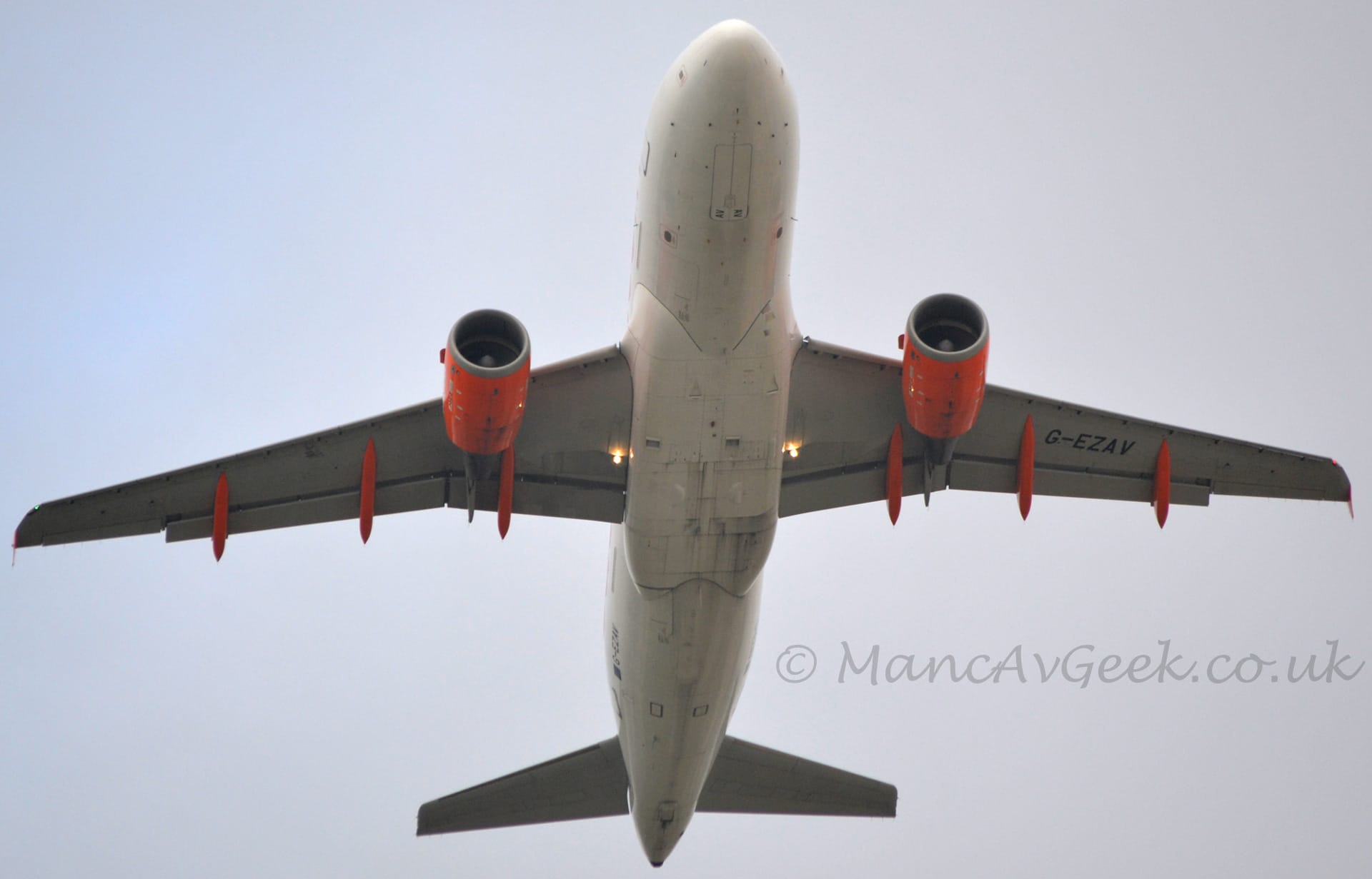 View from underneath of a twin engined jet airliner flying almost directly overhead at a low altitude, with flaps extended from the rear of the wings, undercarriage already tucked away, and the nose raised, suggesting it has just taken off. The plane is almost entirely white, with bright orange engines mounted under the wings, as well as bright orange supports connecting the flaps to the rest of the wing, with 3 on each side. The black registration "G-EZAV" is under the left wing, out-board of the engine pod, between the 2 outer flap supports. Hazy grey sky fills the rest of the frame.