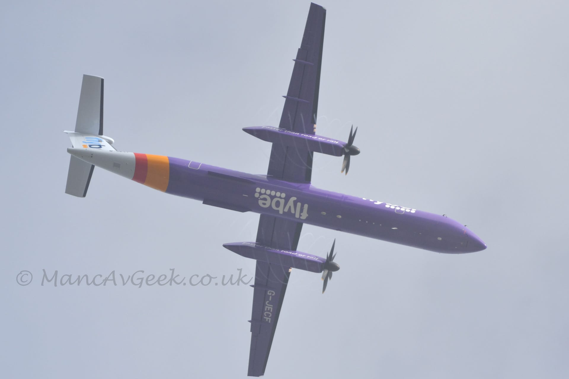 View from pretty much underneath of a high-winged, twin propellor-engined airliner flying from left to right at a low altitude, with flaps extended from the rear of the wings and undercarriage already retracted, suggesting it has just taken off. The plane is mostly extremely purple, with a wide yellow and slightly narrower red band around the rear fuselage, and a white tail and tail-cone. There are white "Flybe" titles over 6 white dots on the forward fuselage, repeated under the belly, and white text "Faster than road or rail" on the sides of the engines under the wings. The white registration "G-JECF" is on the bottom of the left wing. There are blue "Flybe" Titles on the tail. Spinning vapour trails are visible around the engine pods, created by the propellor blades spinning through damp air. Grey sky fills the rest of the frame.