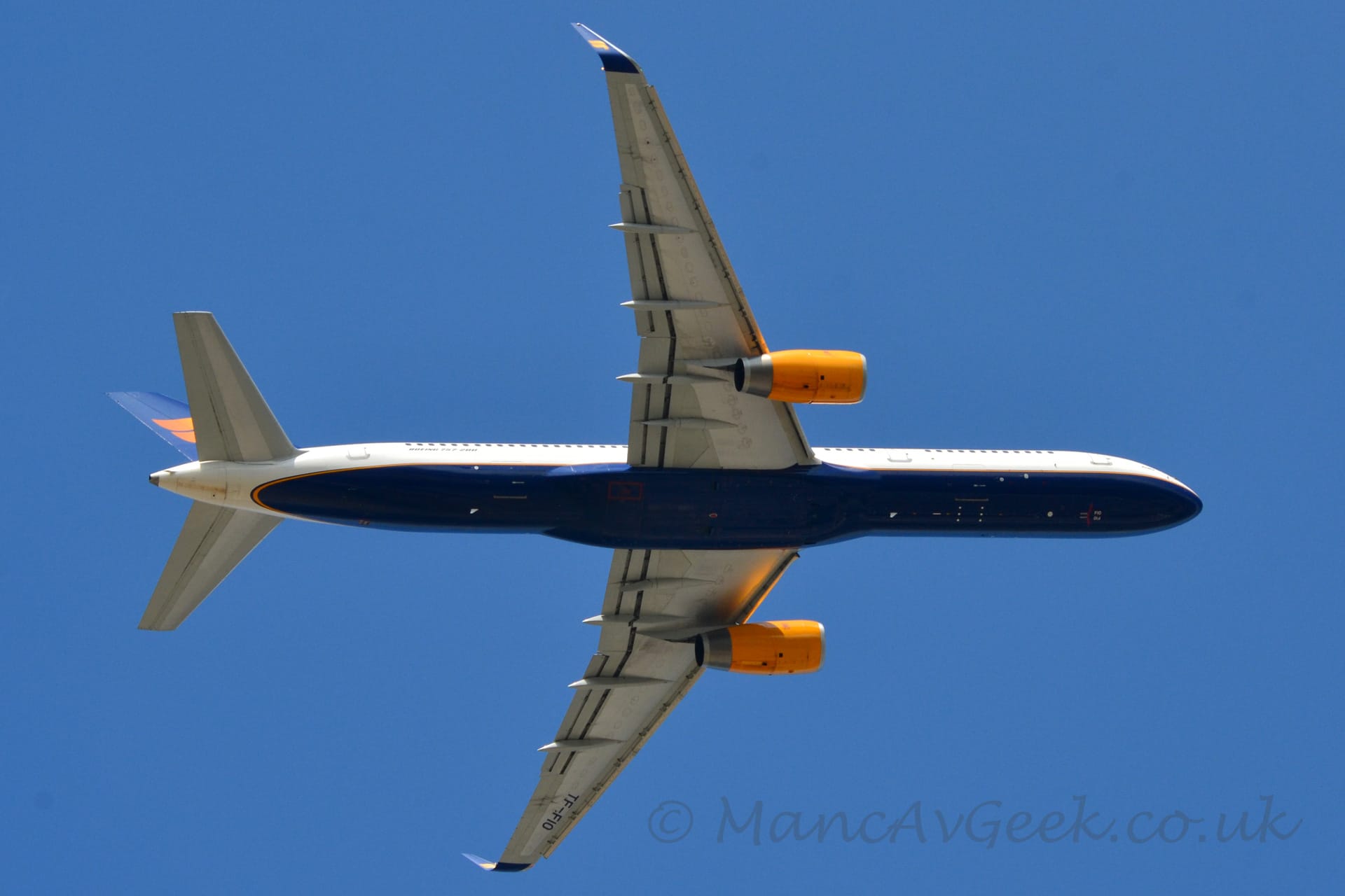 View from underneath of a twin engined jet airliner flying from left to right almost directly overhead, with flaps extended from the front of the wings, undercarriage already tucked away, and the nose raised sharply, suggesting it has just taken off. The plane is mostly white, with a dark blue belly outlined with a yellow pin-stripe. The engines mounted under the wings are a bright yellow. The tail is also dark blue, with a pair of wavy yellow lines in the middle. The up-turned wing-tips are also dark blue with a pair of bright yellow wavy lines. The black registration "TF-FIO" can be seen under the outer sections of the left wing. Vivid blue sky fills the rest of the frame.